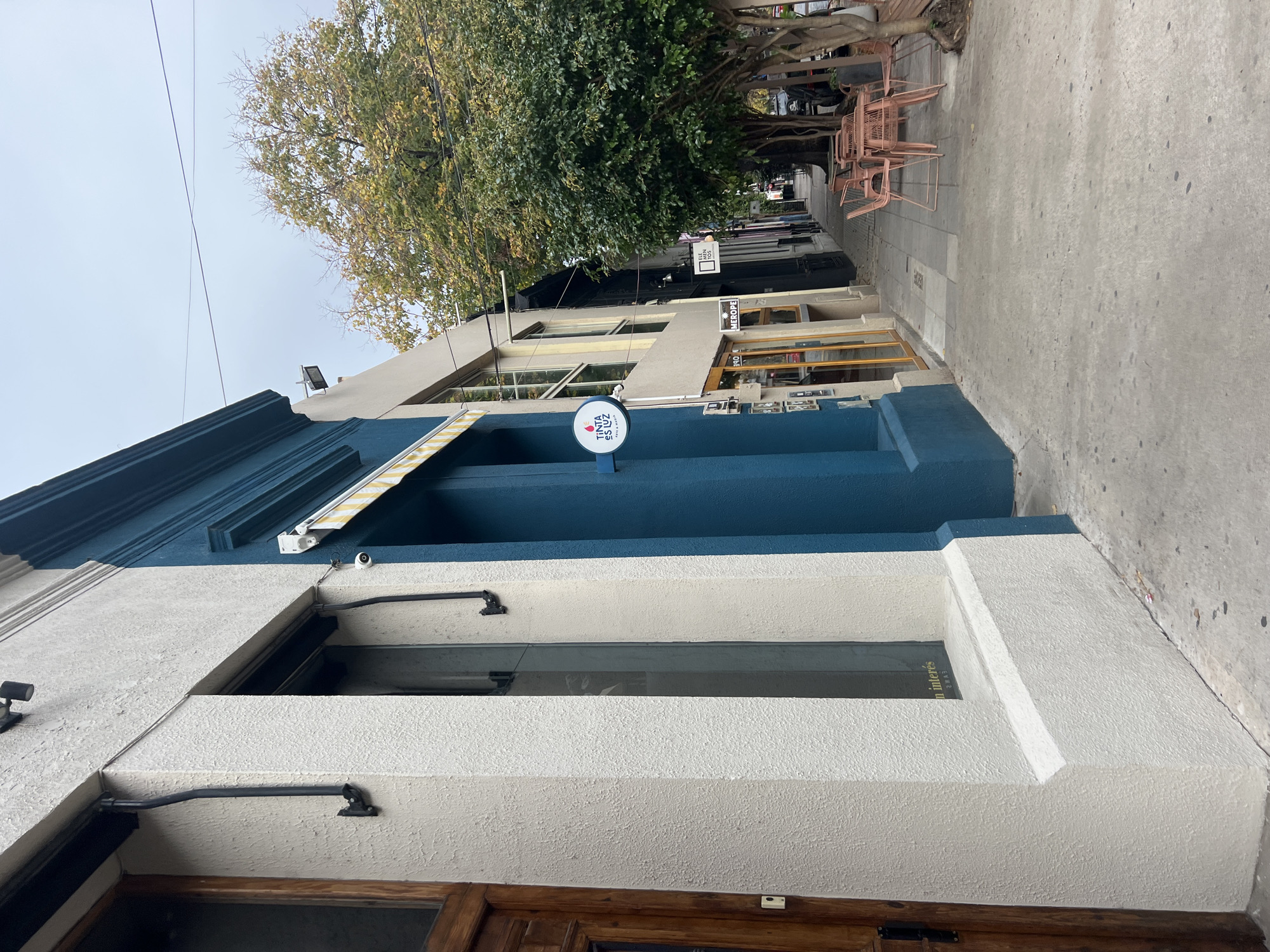 The image shows a building in Buenos Aires, Argentina. The building has a blue and white facade with a sign that says "Tinta es Luz." There is a window above the door with the word "n interés" visible. On the sidewalk, there are several pink chairs and tables, suggesting an outdoor seating area for a cafe or restaurant. A tree with green foliage is visible, partially obscuring the view further down the street. The sky is overcast.