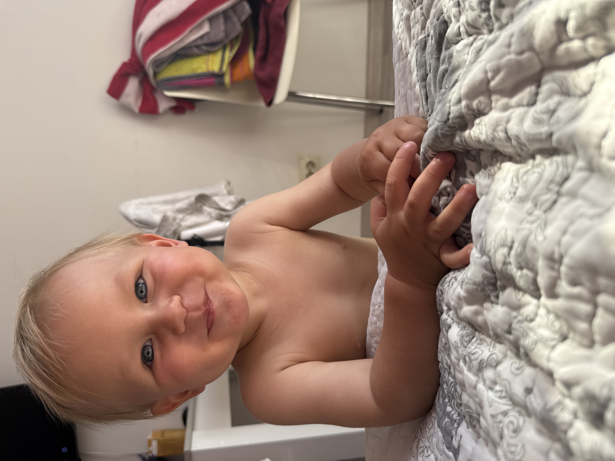 The media file shows a close-up, rotated view of a young child, likely a toddler, lying down. The child has fair hair and blue eyes, and is smiling at the camera. Their torso is bare, and they are partially covered by a textured white and grey blanket. The child's hands are visible, resting on the blanket.
In the background, a shelf with folded clothes is visible, including items with red and white stripes. To the left, a white surface and what appears to be a black object are partially visible. A power outlet is also visible on the wall in the background. The scene appears to be indoors, likely in a bedroom or living area. The lighting suggests it is daytime. There is no visible text in the image. The overall emotion conveyed is cheerful and innocent due to the child's smile. The location context is Sochi, Russia.