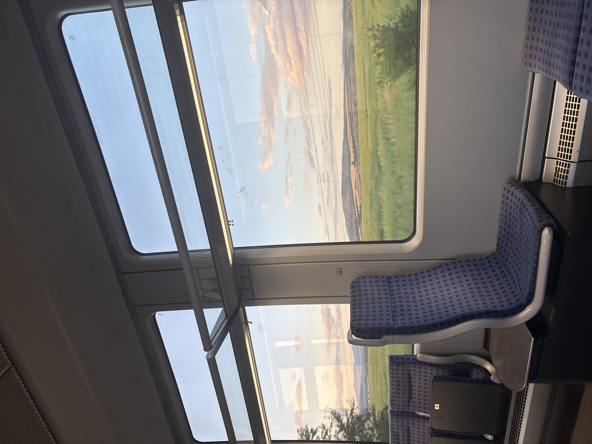 The image depicts the interior of a passenger train car, looking out through its windows at a rural landscape during what appears to be late afternoon or early evening. The scene is described as taking place in Tsar Asen, Bulgaria.
No individuals are present in the train car. The interior features a light gray ceiling, with a textured, possibly ribbed, section on the upper left and a smooth surface dominating the rest. Linear artificial light fixtures are active along the ceiling, casting subtle reflections onto the windows.
Multiple large windows frame the exterior view. On the left side, two vertically arranged windows show a partial, slightly blurred view of a green tree close to the train, with distant rolling hills and a soft, cloudy sky illuminated by warm, golden light. Reflections of the train's interior, including the ceiling and light fixtures, are prominent in these windows.
The primary exterior view is through a large, rectangular window on the right, which dominates the frame. This window overlooks an expansive, vibrant green agricultural field in the immediate foreground and midground. Beyond the field, a series of softer, undulating hills recede into the distance under a pale blue sky streaked with diffuse pink, orange, and white clouds, characteristic of sunset. Several parallel overhead power lines or communication lines are visible crossing horizontally across the sky in the upper portion of the exterior view. A small, dark bush or clump of trees is visible in the lower right corner of the field.
The train's interior is furnished with blue passenger seats featuring a small, repeating grid pattern. These seats have light gray, curvilinear frames. A prominent empty seat in the mid-right foreground faces away from the camera, with a small, dark gray fold-down table extended from its back. To the far left, the backs of two other blue seats are partially visible, one featuring a black tray table with the number '9' printed on it. A vertical structural pillar between the right-side windows displays a white sticker with the number '55' in black text. The floor is dark. A metallic grille, likely for heating or ventilation, is situated at the base of the wall below the rightmost window.
The overall illumination combines the warm, fading natural light from outside with the cooler, artificial lighting inside the train, creating a serene and transitional atmosphere.