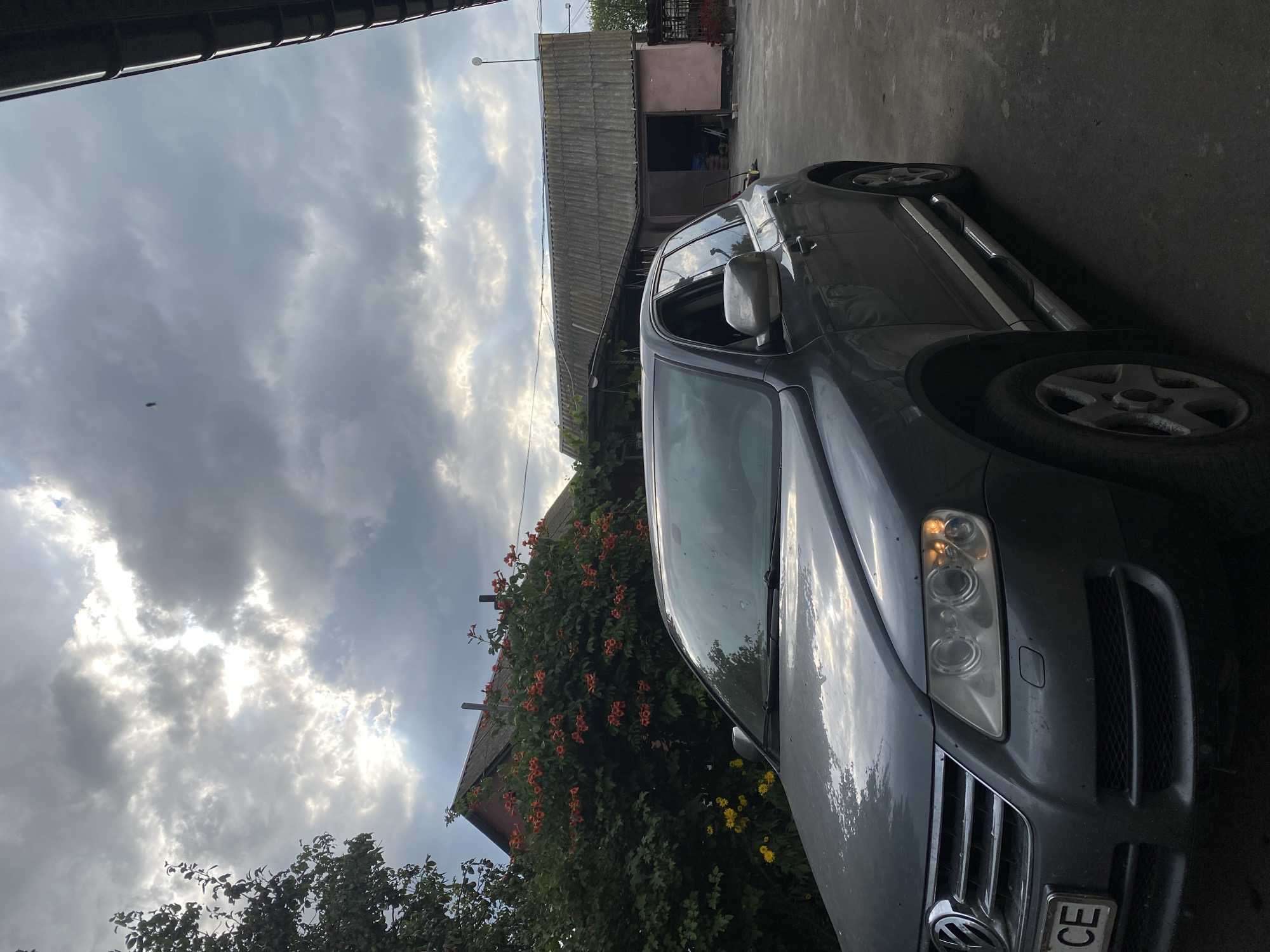 The image captures a gray Volkswagen Touareg parked on a paved surface in front of a building. The building appears to be a workshop or garage with a corrugated roof. Behind the car and to the left, there is a red-roofed structure partially obscured by a dense bush with orange flowers. Yellow flowers are visible below the orange-flowered bush. The sky is overcast with dramatic clouds. A single small, dark object is visible in the sky to the upper right of the frame, possibly a bird or insect. The overall scene suggests a rural or semi-rural setting.