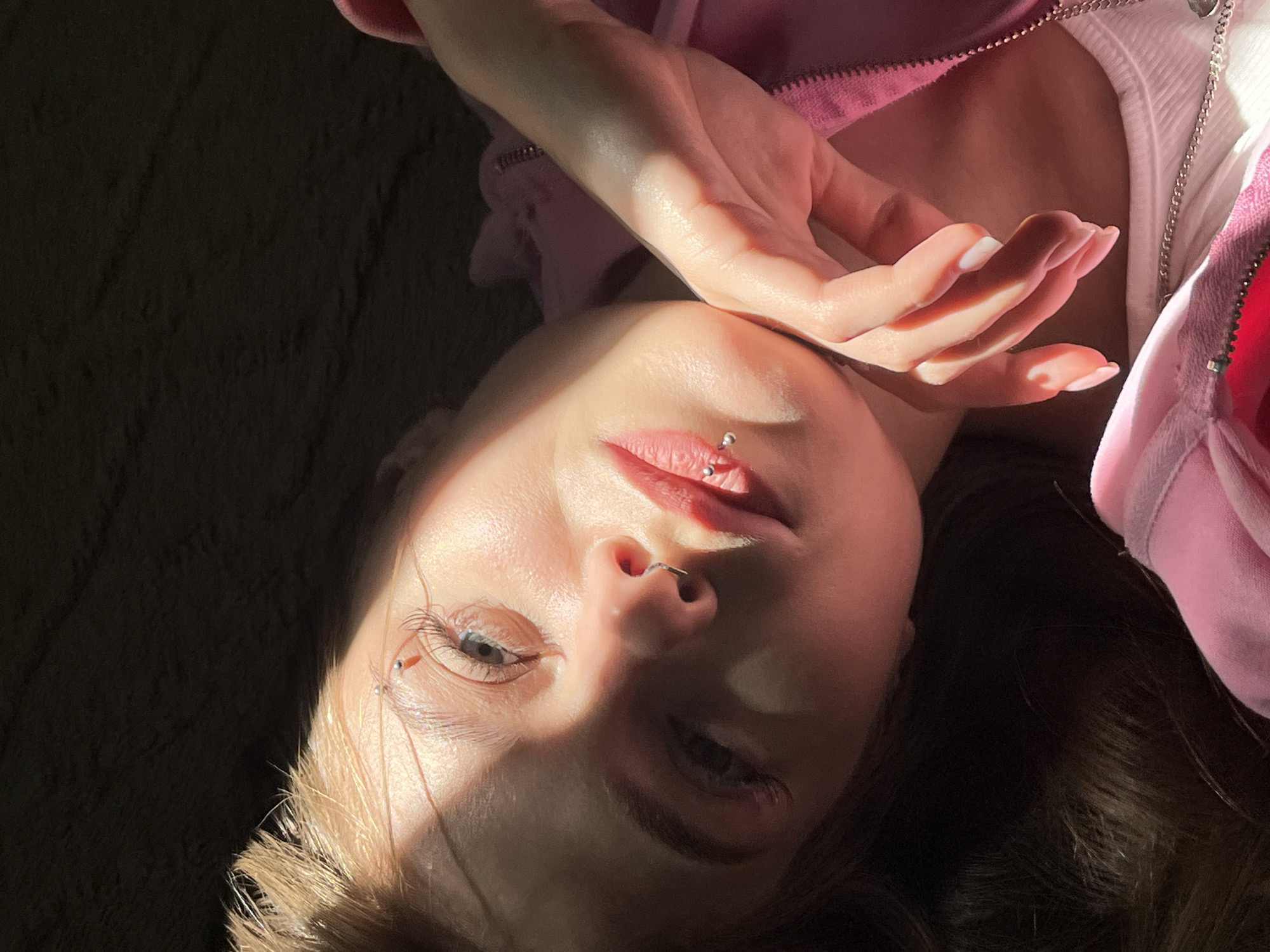 A young woman lies on a dark, possibly textured surface. She is angled towards the camera, with her face illuminated by a strong light source creating stark shadows. Her hair is brown and partially obscures the left side of her face. She has fair skin, and visible piercings on her nose and lower lip. Her right hand is raised slightly above her face. She is wearing a pink hooded jacket over a white top. The exact location is indeterminable from the given image. There is no information available regarding an association with the city of Фоки, Russia.