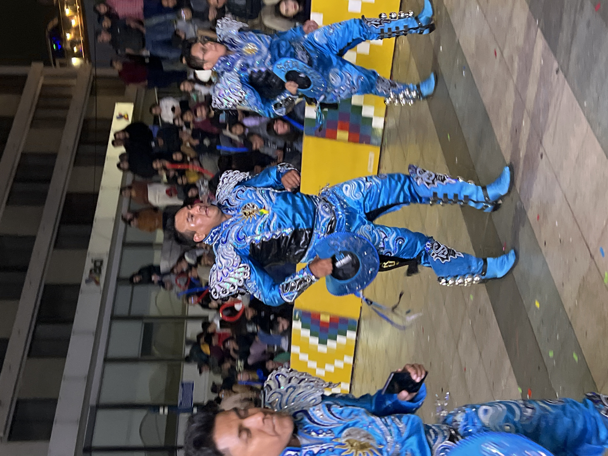 The image depicts a nighttime street performance in Arica, Chile, likely a parade or festival. Three individuals are prominently featured, dressed in elaborate, matching blue costumes with silver and white embellishments. These costumes include intricate patterns on the suits, padded shoulder pieces, and tall, buckled boots. The central figure is holding a blue hat. The performers appear to be in motion, suggesting a dance or procession.
Behind the performers, a crowd of spectators is visible, many of whom are looking towards the dancers. The background includes a building with a sign that appears to read "Adi," and the facade has large windows. A yellow barrier with a colorful, pixelated pattern is situated behind the central and rightmost performers. The ground is paved and strewn with small pieces of colorful confetti. The overall atmosphere suggests a lively cultural event.