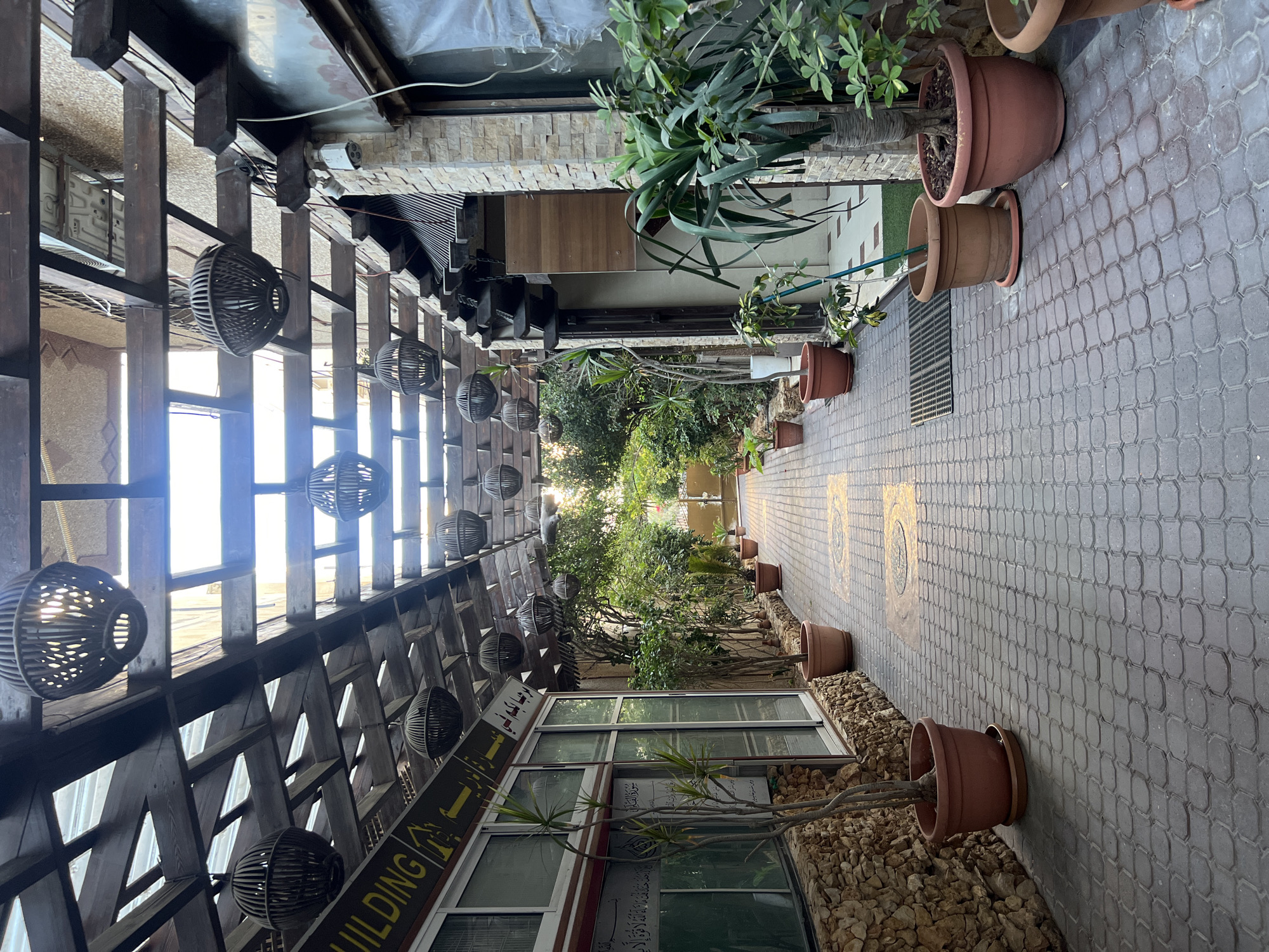 The image shows a covered outdoor walkway in Gaza City, Palestinian Territories. The walkway is paved with interlocking bricks and lined with potted plants. A wooden trellis with hanging spherical lanterns covers the walkway. On the left, there is a building with a stone facade and large windows displaying Arabic text. On the right, a building with a stone facade features a closed door. The scene appears to be an entrance or passage to a commercial or residential area. The lighting suggests it is daytime.