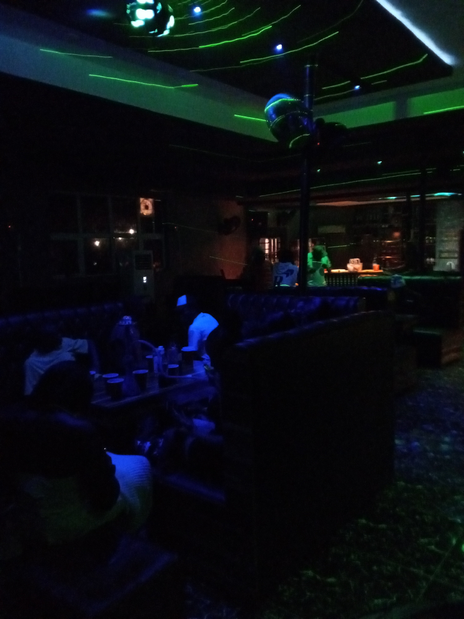 An indoor dimly lit club environment. The scene is dominated by dark tones, with intermittent flashes of neon green lights crisscrossing the ceiling and walls. A disco ball, illuminated with blue and green light, hangs from the ceiling, casting an atmospheric glow. In the foreground, dark-colored plush seating lines the perimeter of the room. Several individuals are seated at tables, some with drinks. A pole is visible in the center of the room, suggesting a dance or performance area. The flooring appears to be carpeted and reflects some of the ambient lighting. The overall impression is of a lively nightlife venue, likely a nightclub or lounge.