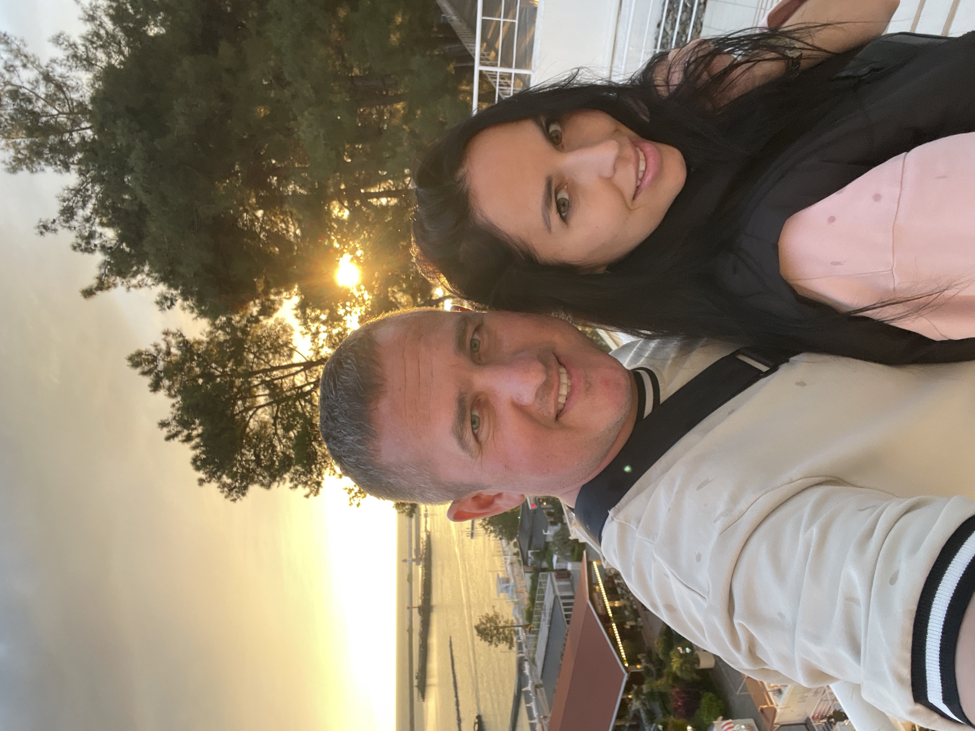 A couple poses for a selfie with a sunset backdrop, likely in Sochi, Russia, judging by the presence of palm trees and coastal architecture. The man has short, graying hair and wears a light-colored shirt with black and white striped trim on the sleeve. He's smiling at the camera. The woman has long, dark hair and is wearing a black top over a pink garment. She is positioned behind and slightly above the man, also smiling at the camera. In the background, there are trees, water, and buildings, all illuminated by the setting sun. The lighting suggests it is either sunrise or sunset. The picture is oriented so that the people are on the right side of the image, with the cityscape on the left.