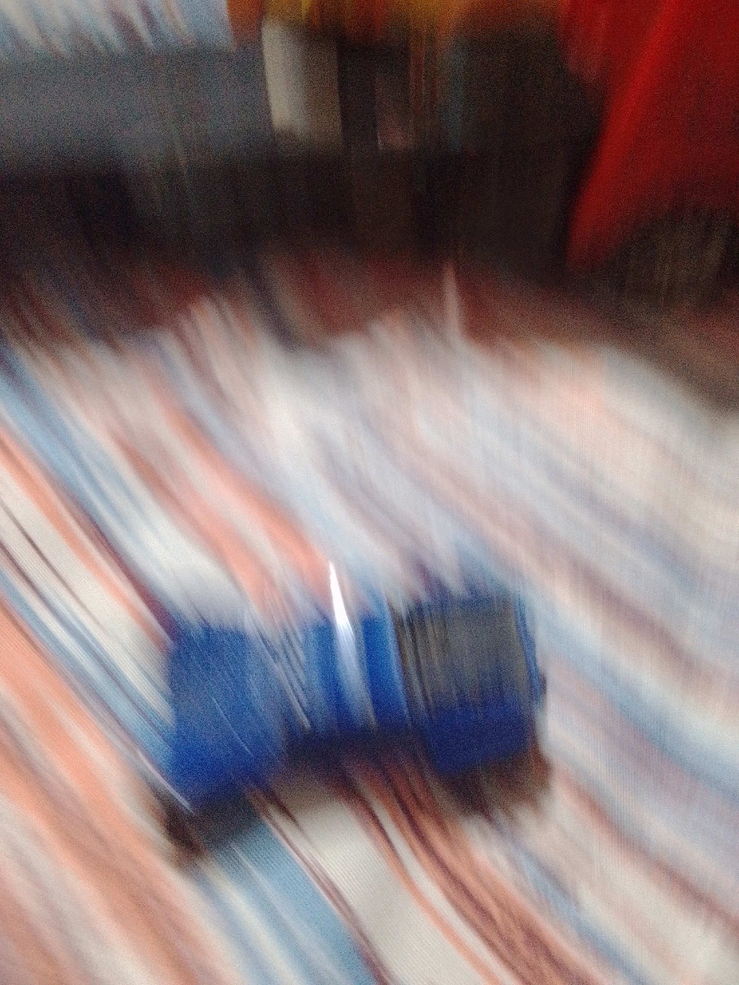 The image is a close-up, motion-blurred photograph of a toy truck. The truck is predominantly blue with some darker, possibly black, sections. It appears to be a toy replica of a larger vehicle, perhaps a transport truck. The background is a fabric with a pattern of diagonal stripes in colors such as white, light blue, and a peachy or light orange hue. The blur suggests movement, either of the camera or the toy itself during the capture of the image. The lighting is not clearly discernible due to the blur, but it appears to be indoor lighting. There is no visible text in the image. The context provided, La Tagua, Venezuela, does not offer any specific visual cues within the image itself.