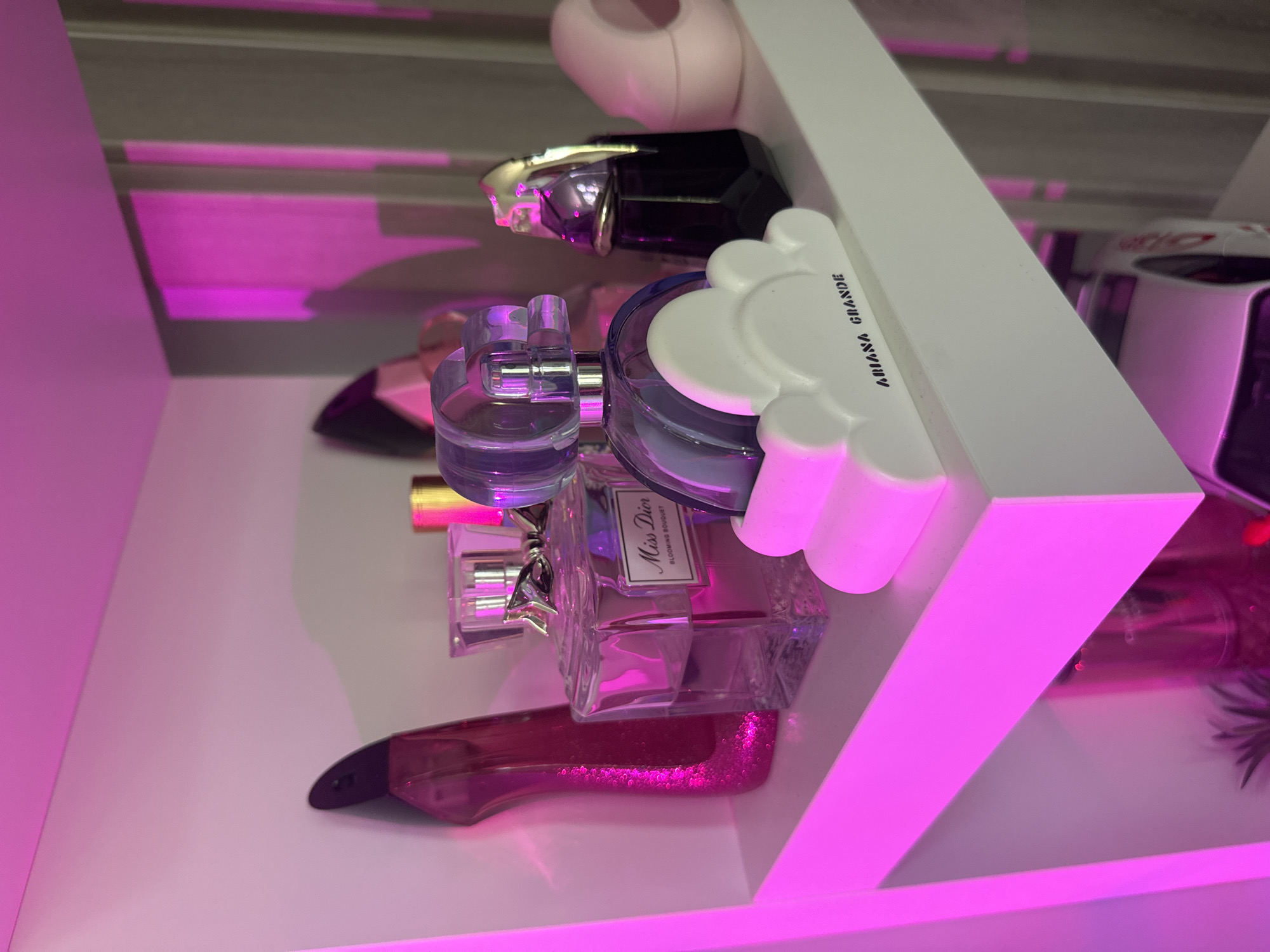 The image depicts a white, illuminated shelf in an indoor setting. The primary light source bathes the scene in a strong magenta-pink hue, casting colored reflections and shadows across the objects and surrounding surfaces.
Arranged on the shelf, from left to right, are several perfume bottles:
1. On the far left, a tall, high-heeled shoe-shaped bottle, dark pink or magenta, with a glittery texture.
2. Next to it, a rectangular glass bottle with a silver top and a silver bow around its neck. A label on the front clearly reads "Miss Dior" and "BLOOMING BOUQUET." The liquid inside is a translucent pinkish-purple.
3. In the center, a translucent purple spherical perfume bottle rests on a white, cloud-shaped base. The base is inscribed with "ARIANA GRANDE" in black text.
4. To its right, a dark, possibly black or deep purple, bottle with a gold-colored stiletto heel forming its cap.
5. On the far right, a light pink, rounded, abstractly shaped object with a central cavity, possibly another perfume bottle or decorative item.
The background consists of a wall, appearing to be a light neutral color, heavily affected by the pink lighting, showing faint vertical lines suggesting a blind or curtain texture in the distance. Below the main shelf, a glimpse of another white surface is visible, along with part of a white object, possibly a toy car or model. No individuals are present in the image, and no actions are depicted.
