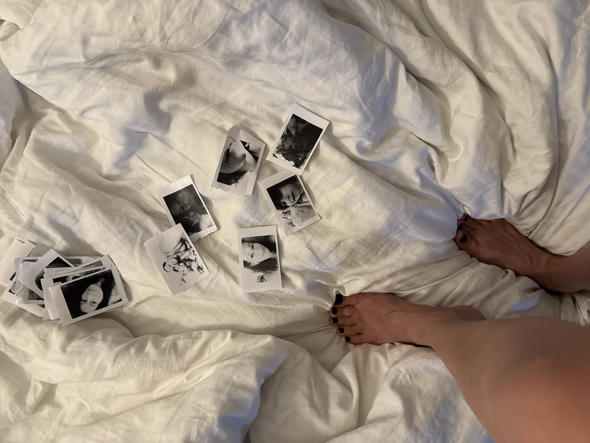 A high-angle shot depicts a bed with rumpled white sheets. Scattered on the bed are several black and white instant photos. In the lower right corner, a person's feet with black toenail polish extend from under the sheets.
The scene is unlikely to be in Chaikovsky, Russia, given the details present do not imply such a location. The photograph captures a domestic interior scene centered on the bed, photographs, and feet.