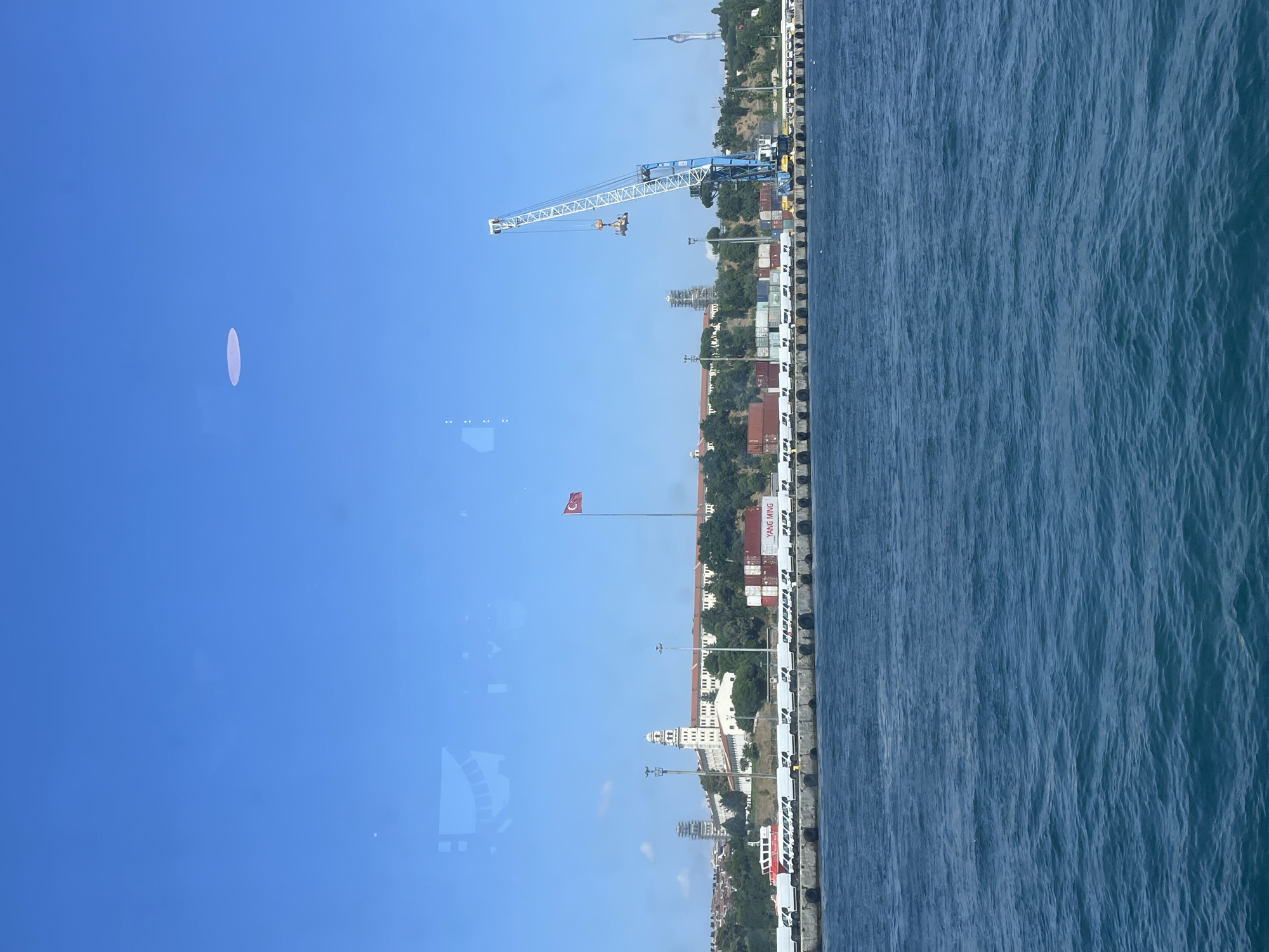 This is a photograph of a coastal scene in Istanbul, Turkey, taken from a body of water. In the foreground, a large expanse of blue, slightly rippled water fills the lower half of the image. Beyond the water, a long, grey concrete quay stretches horizontally across the middle ground. Lined up along the quay are numerous white delivery vans. Behind the vans, stacked shipping containers, some red and some white, are visible. A large blue and white gantry crane is positioned on the quay, with its arm extended over the containers, lifting a load.
In the background, a long, low-rise white building with a distinctive tower stands prominently on a hill, bordered by lush green trees. A Turkish flag flies from a flagpole in front of this building. To the left of this building, a red and white vessel is docked at the quay. Further in the distance, the cityscape of Istanbul is hinted at with more buildings and structures, including a tall, slender tower on the right side of the frame. The sky is a clear, bright blue. The overall impression is that of a busy port area under a sunny sky.