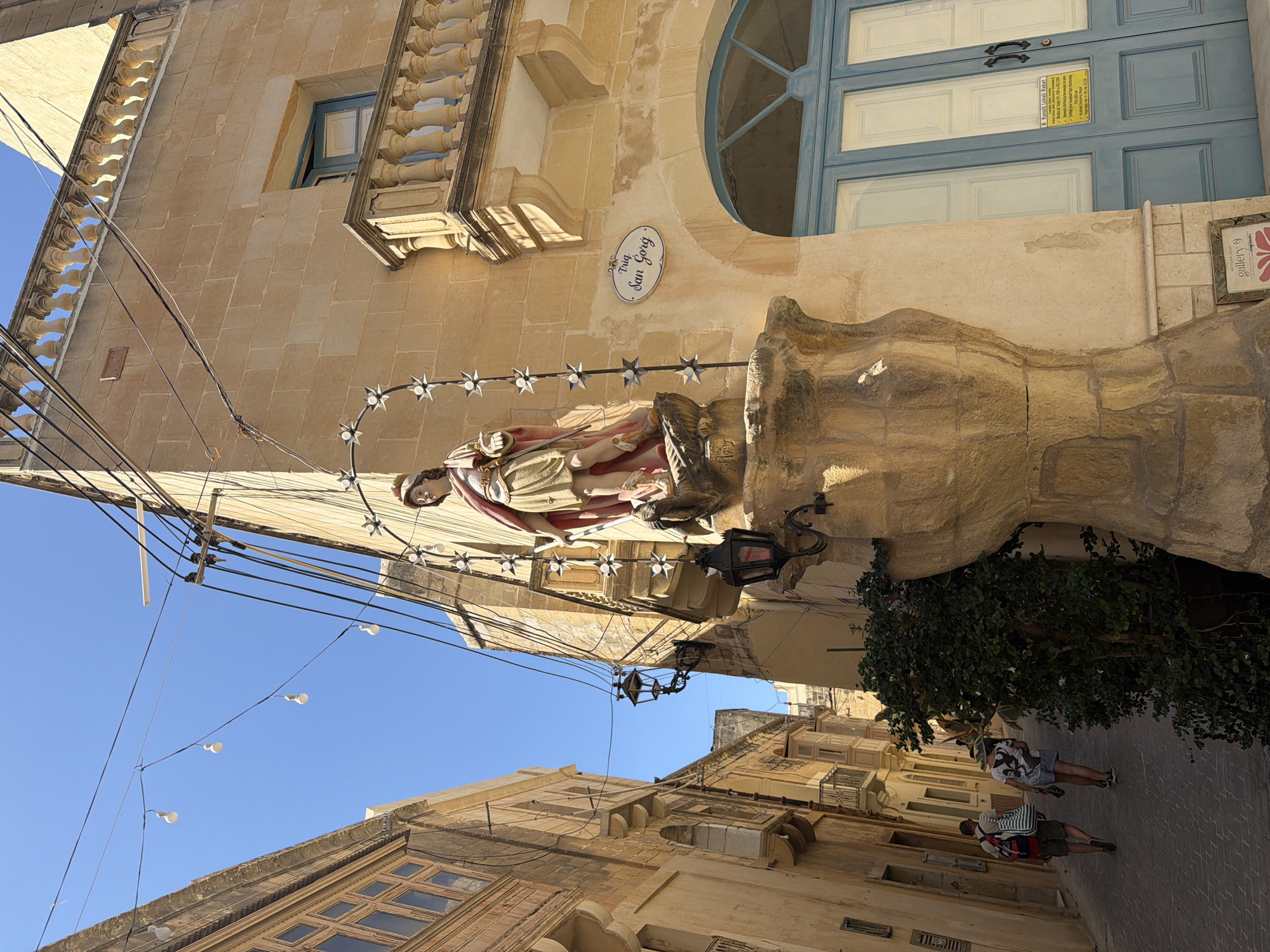 The image depicts a narrow, paved street in Victoria, Gozo, Malta, likely in the historic center. The street is lined with traditional Maltese limestone buildings, characterized by their warm, sandy color and architectural details such as balconies and decorative stonework.
Dominating the foreground is a religious statue, possibly Saint George, mounted on a stone pedestal. The statue appears to be made of painted stone or plaster and is adorned with a red cloak. The pedestal itself is intricately carved and weathered. Above the statue, a string of decorative lights with star-shaped bulbs is strung across the street, along with electrical wires and street lamps.
To the right of the statue, a building features a balcony with balustrades and a blue-painted door. A sign on the wall near the door reads "Trig. San Gorg," identifying the street as Triq San Gorg (Saint George Street).
Further down the street, two individuals are walking away from the camera. One person, on the left, wears a backpack and shorts, carrying a striped tote bag. The other person, on the right, wears a t-shirt and shorts. Their presence suggests this is a public thoroughfare.
The sky is clear and blue, indicating a sunny day. Lush green foliage is visible at the base of the statue pedestal and also further down the street, adding a touch of nature to the urban landscape. The overall impression is one of a charming and historic Mediterranean town street scene.