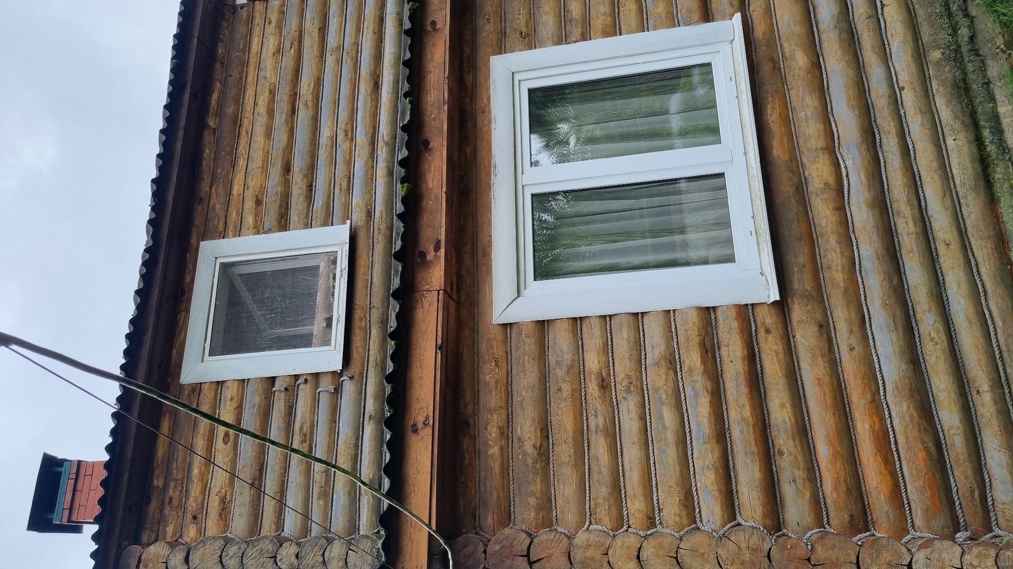 The image shows the exterior wall of a log cabin in Солнечная Поляна-3 Снт, Russia. The wall is constructed from horizontal logs, with rope detailing filling the gaps between them. There are two white-framed windows visible on the wall. The upper window is smaller and appears to have a screen. The lower window is larger and is covered by light-colored curtains. Above the windows, a corrugated roofline is visible, and a brick chimney with a black cap protrudes from the roof. A power line runs diagonally across the upper portion of the image. The overall impression is of a rustic, rural dwelling.