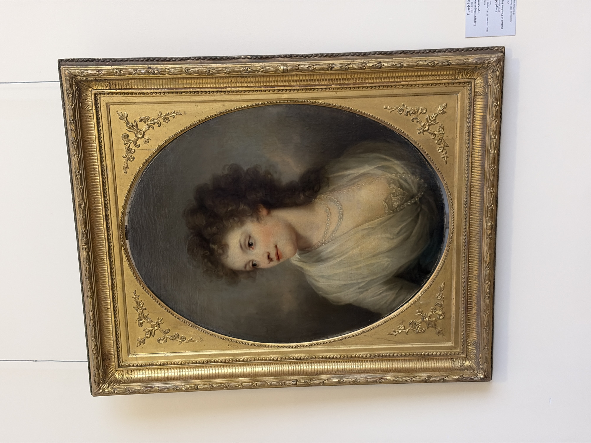 The image shows a framed oval portrait of a woman with dark curly hair and a white dress or shawl. The portrait hangs on a white wall. A label with text in both Russian and English is affixed above the top right corner of the frame. The text on the label identifies the painting as "Portrait of Charlotte, Sophia" by Joseph M. Grassi and notes it was acquired in 1929. While the location isn't explicitly stated in the provided text, the Cyrillic text on the label suggests the artwork is located in Saint Petersburg, Russia.