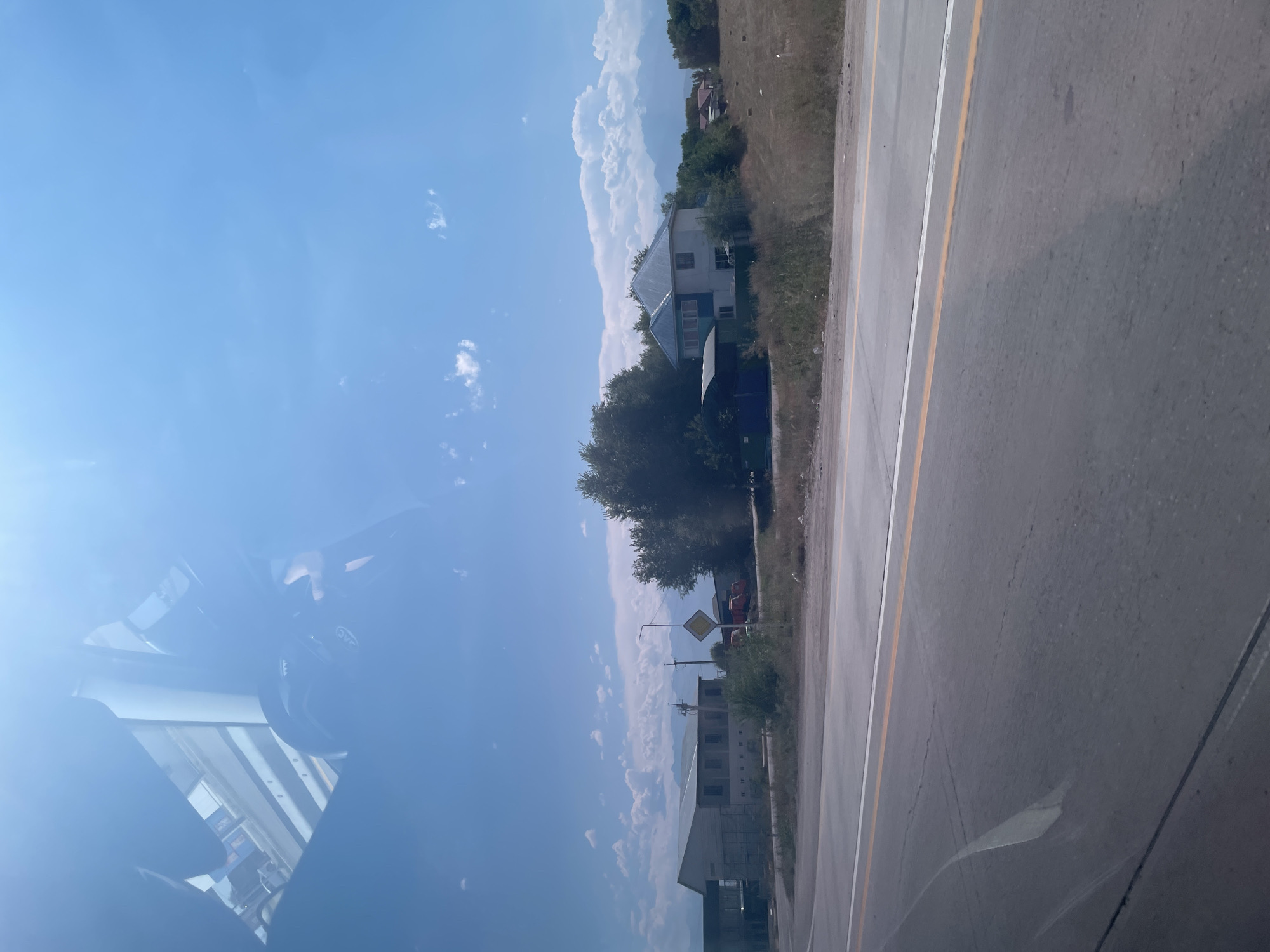 The image shows a roadside scene under a bright, partly cloudy sky. The foreground is dominated by a paved road with yellow and white lane markings, appearing to be a highway or main thoroughfare. The road surface shows some wear and cracking.

To the right of the road, a dry, grassy embankment slopes upward. In the middle ground, several structures are visible. On the left, there is a long, light-colored building with multiple windows and a flat roof, possibly an industrial or commercial building. Further to the right, closer to the road, is a yellow diamond-shaped sign with a black border and symbol, indicating a warning. Behind the sign are several red vehicles, possibly trucks.

Dominating the right side of the middle ground is a two-story house with a light-colored facade, a dark metal roof, and a porch area. The house is surrounded by trees and vegetation. In the background, there are more trees, hills, and distant mountains, suggesting a semi-rural or mountainous environment. The sky is a vibrant blue with scattered white clouds. The reflection of a car's interior, including a dashboard and a hand, is visible on the upper portion of the image, indicating the photo was taken from inside a vehicle. The scene appears to be in or near Tonkeris, Kazakhstan, given the context.