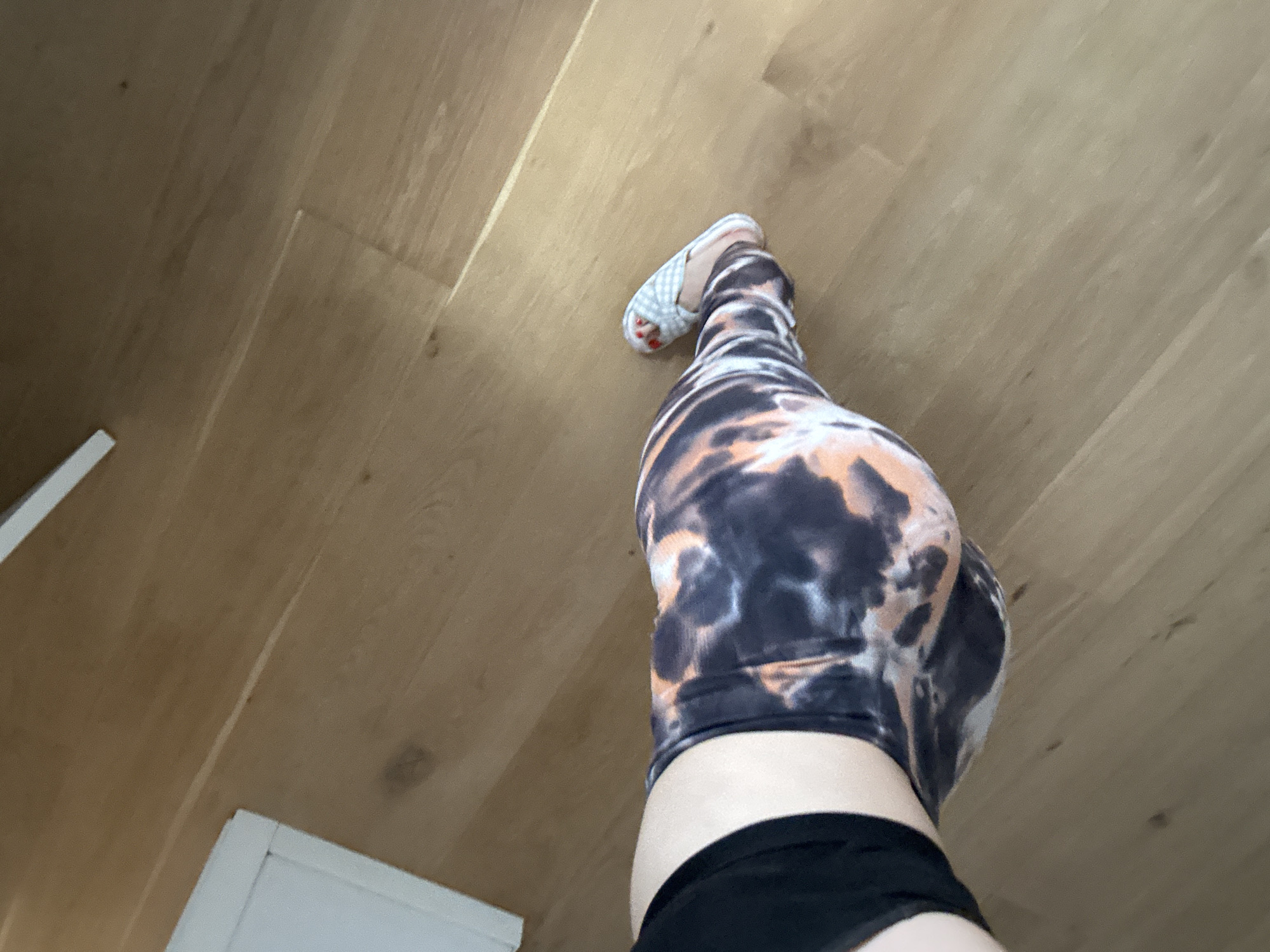 The image shows a close-up, low-angle view of a person's legs and hips wearing tie-dye leggings and checkered slippers. The person is standing on a wooden floor. The lighting suggests an indoor environment. There are no identifiable landmarks or elements that would indicate the location is Murcia, Spain.
