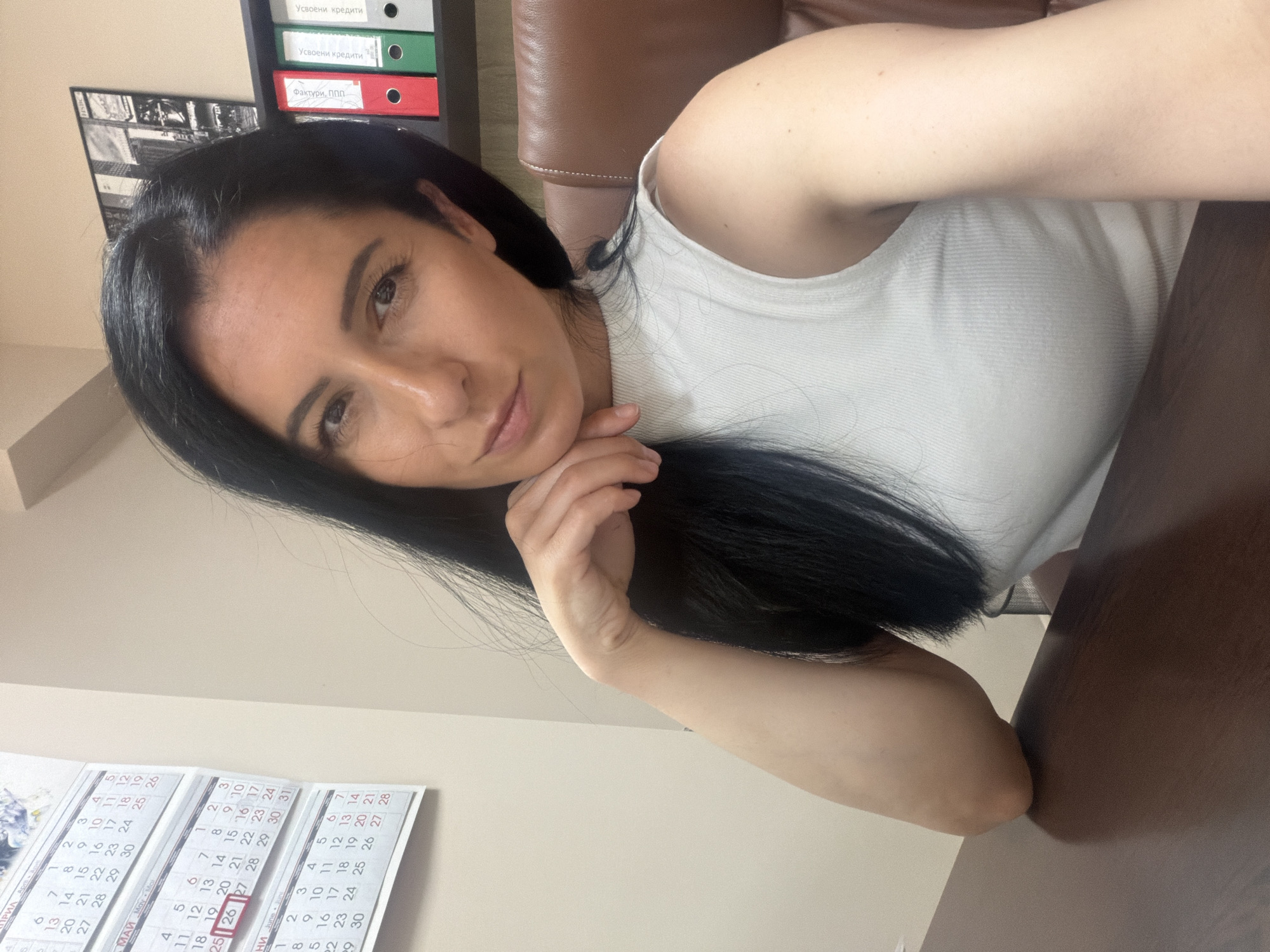 The image is a selfie of a woman indoors. She has long, dark hair and fair skin, and she is wearing a white sleeveless top. Her hand is near her face, seemingly in a thoughtful or relaxed pose.
The background is an office setting. Behind her, there is a shelf with folders, marked in Bulgarian: "Усвоени кредити" (Approved Credits) and "Фактури, ППП" (Invoices, VAT). To the left of the folders, there is a picture or drawing. Below the shelf, there is a calendar. It appears that the calendar is set to May, June, and April. The calendar shows the 26th day of May. The location is Pleven, Bulgaria, based on the language used in the folders. The woman is sitting at a desk with a brown surface. The back of an office chair is partially visible behind her head.