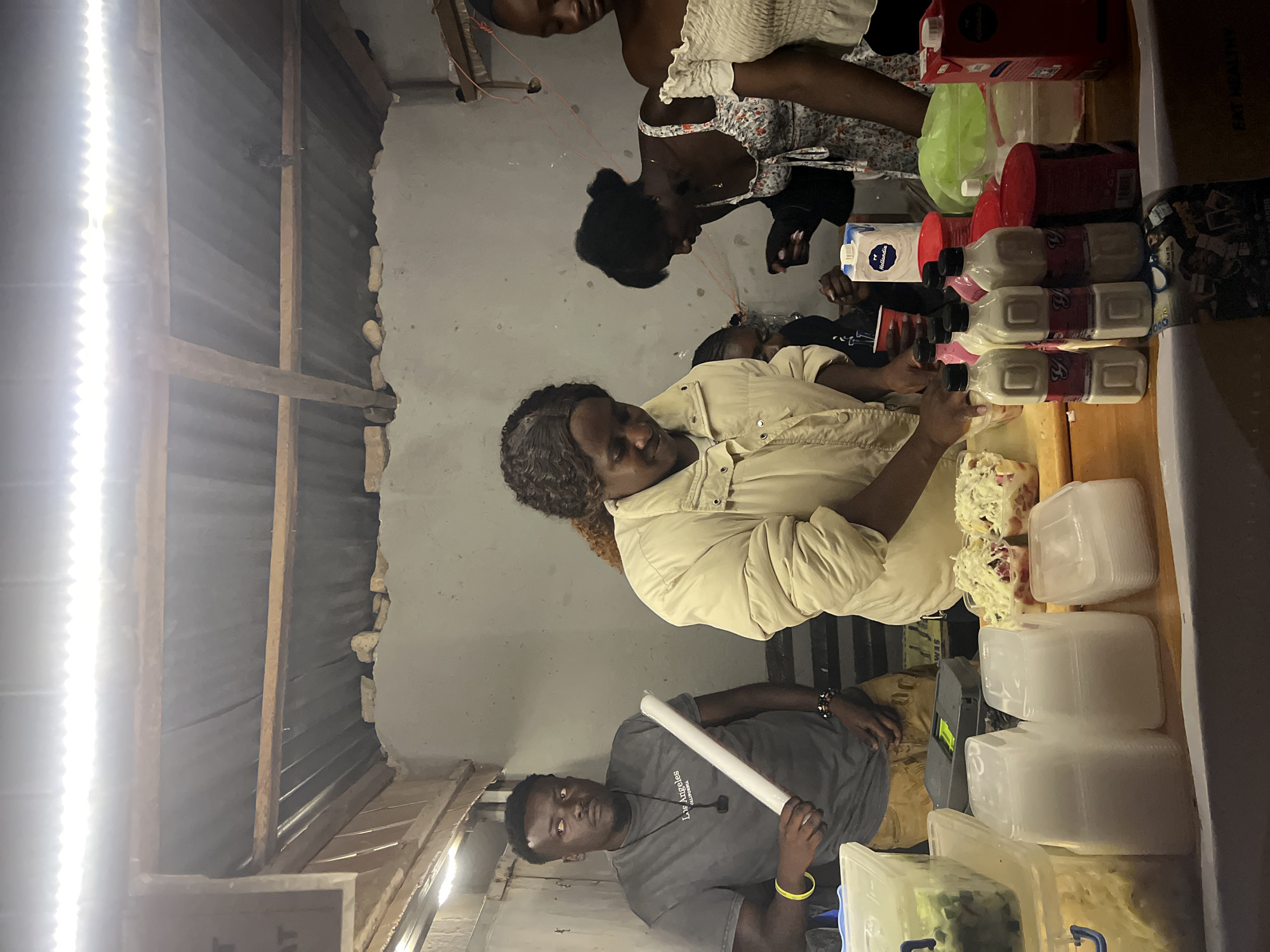 In Durumi, Nigeria, an outdoor food stall is captured at night. A man in a grey t-shirt holding a white cylinder stands to the left. In the center, a woman in a cream-colored puffer jacket prepares food on a wooden counter. Behind her, several other individuals, including two women in patterned dresses and a young girl, are visible. The stall features various food items, containers, drinks, and disposable cups. The background consists of a simple structure with a corrugated metal roof and concrete walls, illuminated by artificial lighting.
