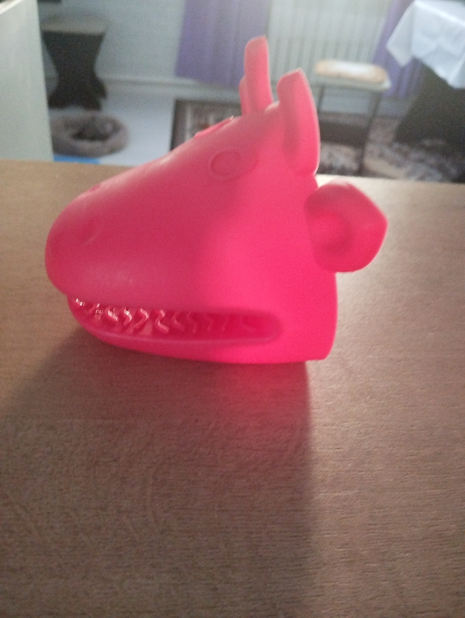 A close-up shot displays a bright pink, plastic toy resembling a stylized animal head, possibly a hippo or similar creature, resting on a brown surface. The surface appears to be a tabletop or countertop. In the blurred background, glimpses of a domestic interior can be seen, including furniture, a pet bed, and a radiator. The location is not discernible from the provided image without additional information.