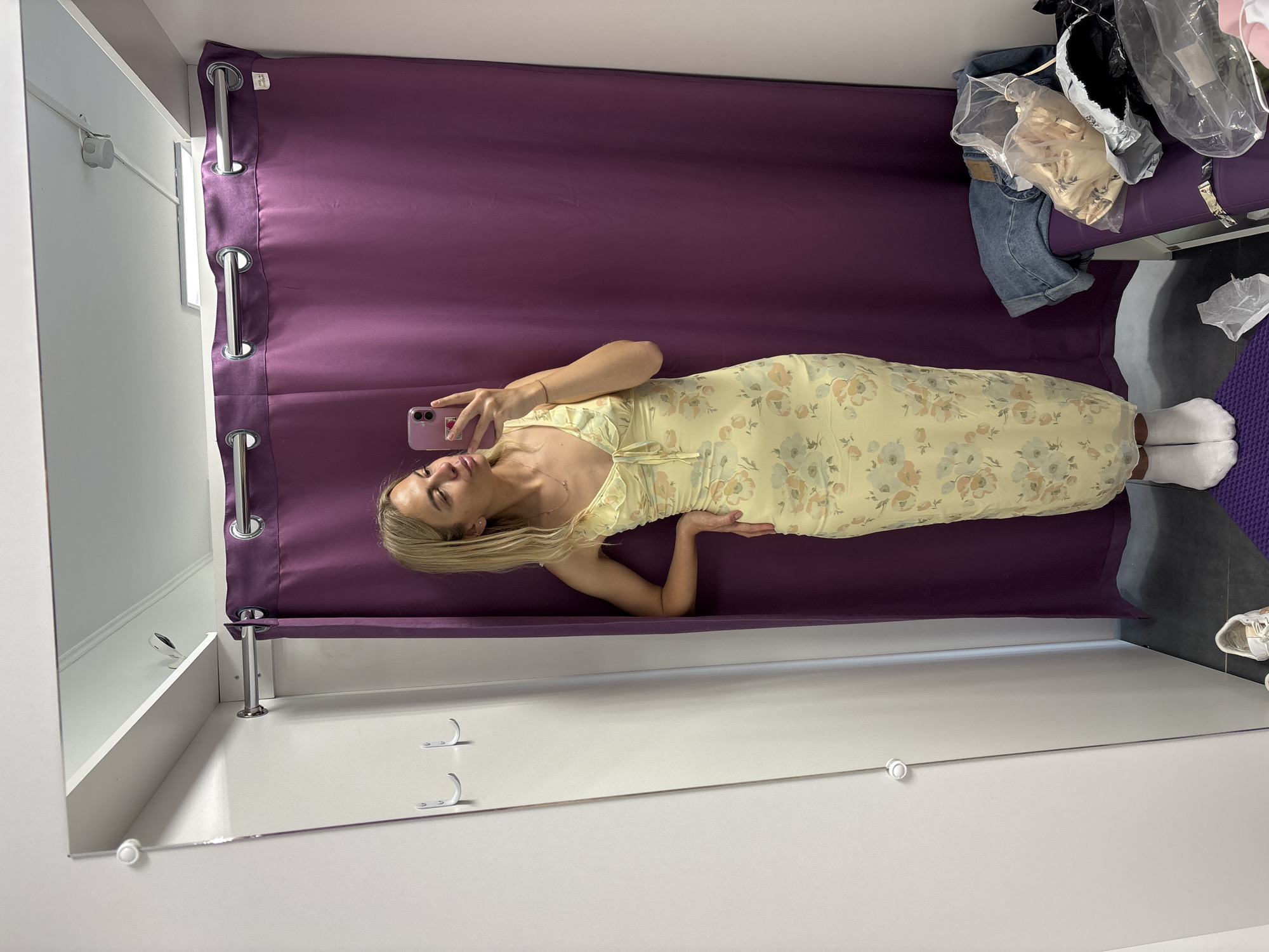 A young Caucasian woman with blonde hair is standing in a fitting room and taking a selfie in a mirror. She is wearing a yellow floral-print slip dress and white socks. The fitting room has a purple curtain, a shelf with a light fixture, and two white hooks on the wall. On a purple bench to the right, there are several items of clothing and bags. The floor is tiled and there is a purple mat under the woman's feet. The overall impression is that she is trying on clothes in a retail store. The image was taken in Krasnoyarsk, Russia.