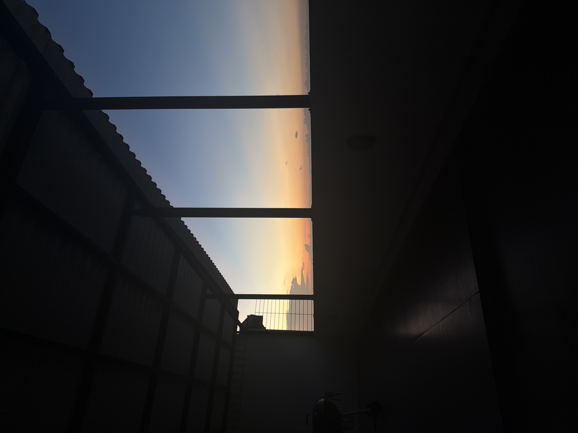 The image shows a view through a window-like opening in a dark room or structure, revealing a sky transitioning from blue to orange. The view is partially obstructed by what appears to be a corrugated metal wall or fence on the left side of the opening. A dark framework divides the view of the sky into sections. The right side of the image is in complete darkness, except for a faint light fixture in the ceiling. The room contains indiscernible objects at its bottom. The city and location are unknown.
