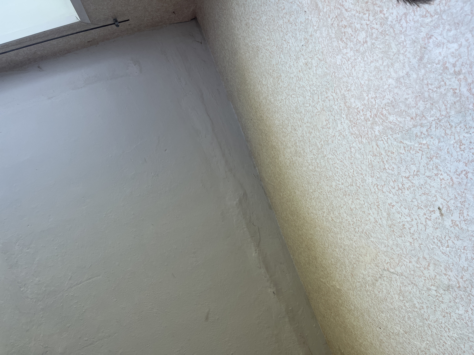 A low-angle, indoor shot shows a textured, light-colored ceiling that appears to be made of plaster or concrete. The ceiling has some subtle imperfections and variations in tone, suggesting age or wear. Below the ceiling, a section of a wall is visible, also light-colored and with a similar textured finish. The foreground of the image is dominated by a floor with a light beige and pink speckled pattern, possibly linoleum or tile. The overall scene suggests an interior space with basic, possibly utilitarian, finishes. There are no people or discernible objects that would indicate a specific location within the city of Krasnye Baki, Russia, beyond it being an indoor environment.