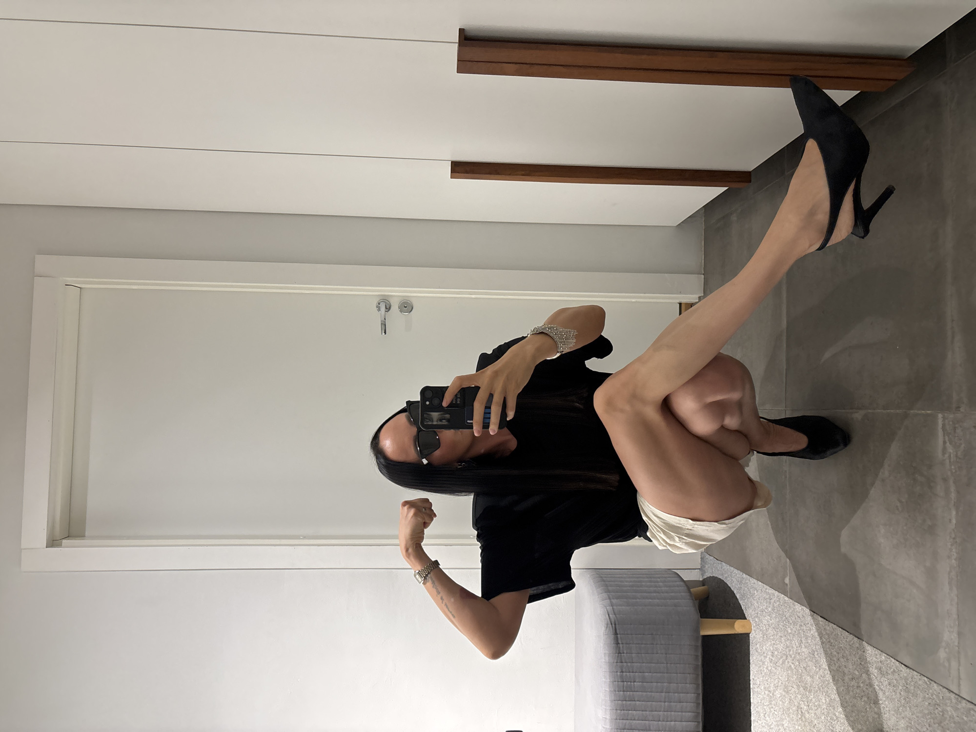 A person is squatting and holding a smartphone to take a selfie in what appears to be an indoor room, likely a bedroom or dressing area. The person is wearing a black top, light-colored shorts, black high-heeled shoes, and sunglasses. They have long, dark hair. Their right arm is raised and flexed. A grey patterned ottoman or bench is situated near the person. The room has a white door, possibly leading to another room or closet, and white walls. There is dark wooden accent on the wall near the ceiling. The floor is covered with large grey tiles. The location in the city of Chaikovsky, Russia is not determinable from the image.