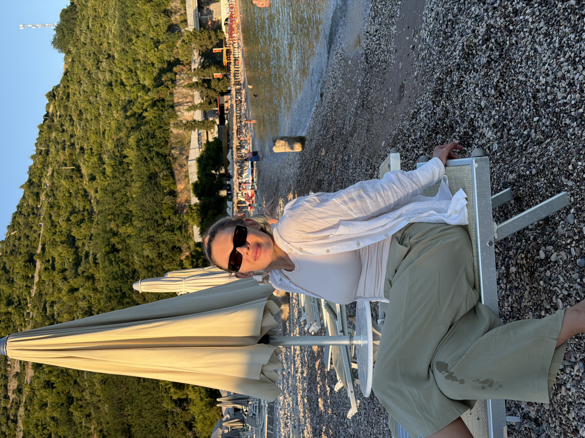 A woman sits on a lounge chair on a pebbled beach in Budva, Montenegro. She is wearing a white unbuttoned shirt over a beige top and loose green pants. The woman has dark sunglasses on and her hair is pulled back. She is smiling and looking at the camera. Behind her, beach umbrellas are set up, and further back, there is a body of water with people swimming and enjoying the beach. The background features a lush green hillside with some buildings nestled into it. The sun is setting, casting a warm glow on the scene.