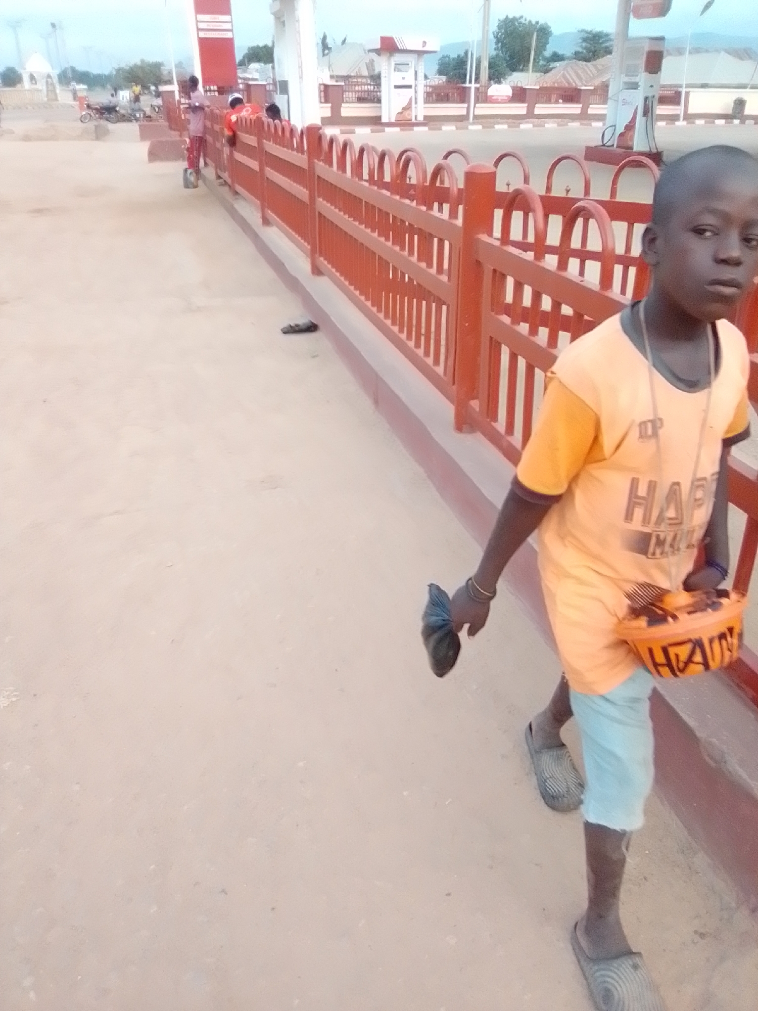 A young boy, likely of Nigerian descent, walks along a dusty path near a gas station in Kwadon, Nigeria. He is wearing an orange t-shirt with a graphic design and light blue shorts. Around his neck, he wears a necklace with an orange container attached, possibly for collecting money or items. In his left hand, he holds a dark-colored object, possibly a shoe or a piece of cloth. He is also wearing grey sandals. The path is bordered by a red wooden fence. In the background, a gas station with pumps and a tall red sign is visible, along with a few buildings and trees. The overall environment appears to be a somewhat arid, outdoor setting.