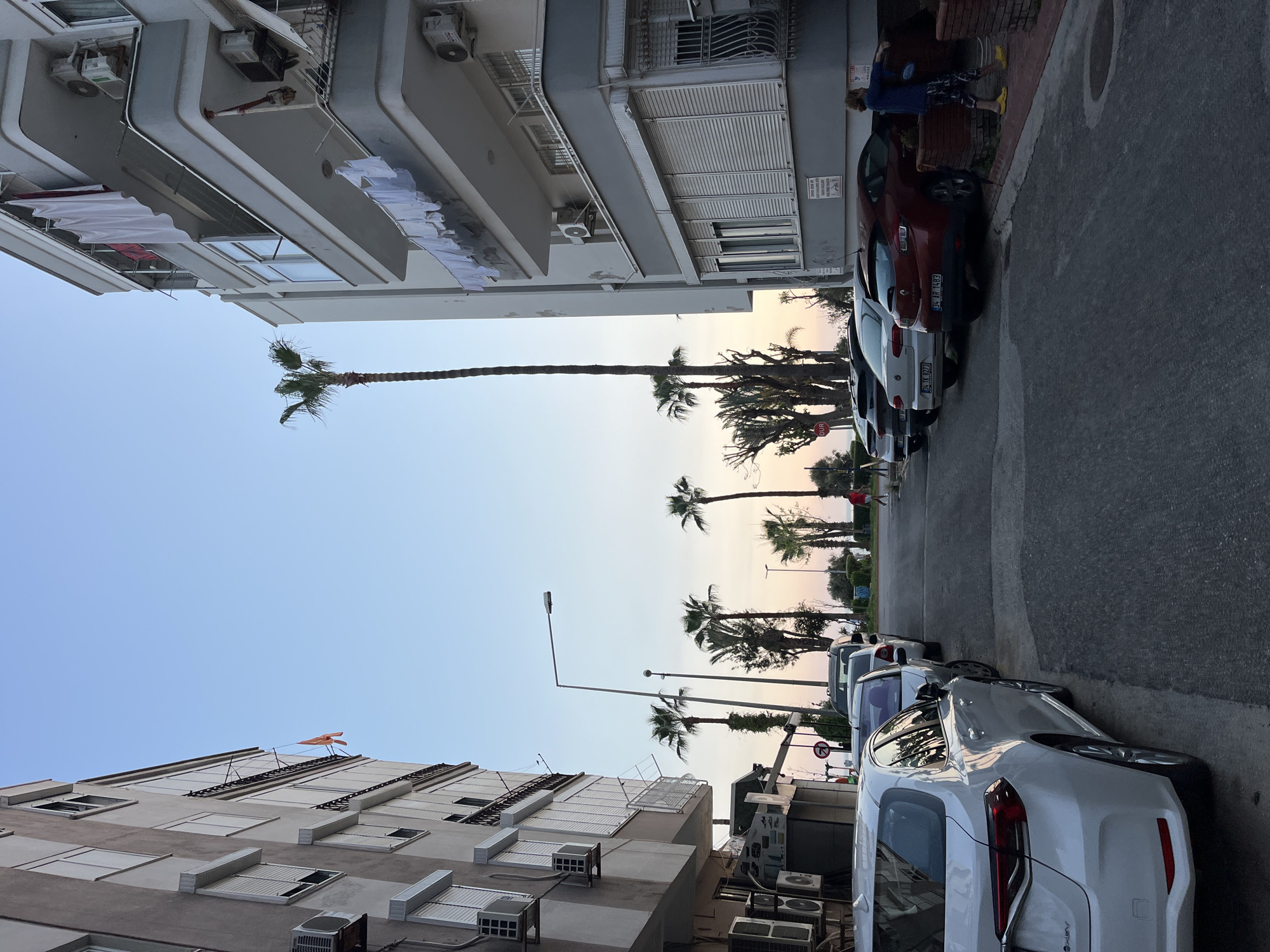 The image depicts a street scene in Alanya, Turkey, under a clear sky displaying a gradient of light blue overhead transitioning to warmer, softer hues near the horizon, suggesting either sunrise or sunset. The street is lined with multi-story buildings and numerous palm trees.
On the left, several buildings feature balconies, windows, and visible air conditioning units. White and red laundry is hung out to dry on multiple balconies. A small red flag, possibly Turkish, is visible atop one of these buildings further down the street.
The asphalt street runs through the scene, with a row of parked cars along the right side. These include a white hatchback (license plate "33 N 0222") and a dark red vehicle (license plate "34 HF 1483"). A prominent red circular "DUR" sign (Turkish for "STOP") is visible near the parked cars. A person wearing a blue top, patterned shorts, and yellow shoes stands on the sidewalk next to the dark red car. Another individual is visible in the distance near the palm trees, closer to what appears to be a waterfront.
Tall palm trees are scattered throughout the landscape, particularly abundant in the middle ground and further in the distance, indicating a coastal or warm climate. Streetlights are also present. The scene conveys a calm, everyday atmosphere of a residential or mixed-use area.
