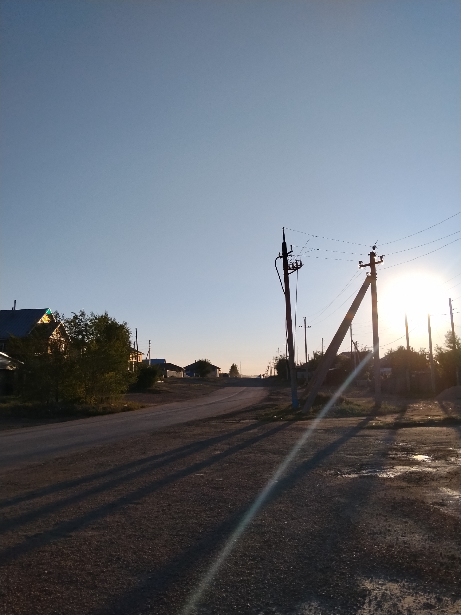 A rural road curves to the left, lined with trees and houses on the left side of the frame. The sun is setting on the right, casting long shadows across the road and the surrounding dirt. Several utility poles with wires are visible along the roadside. The sky is clear and blue.