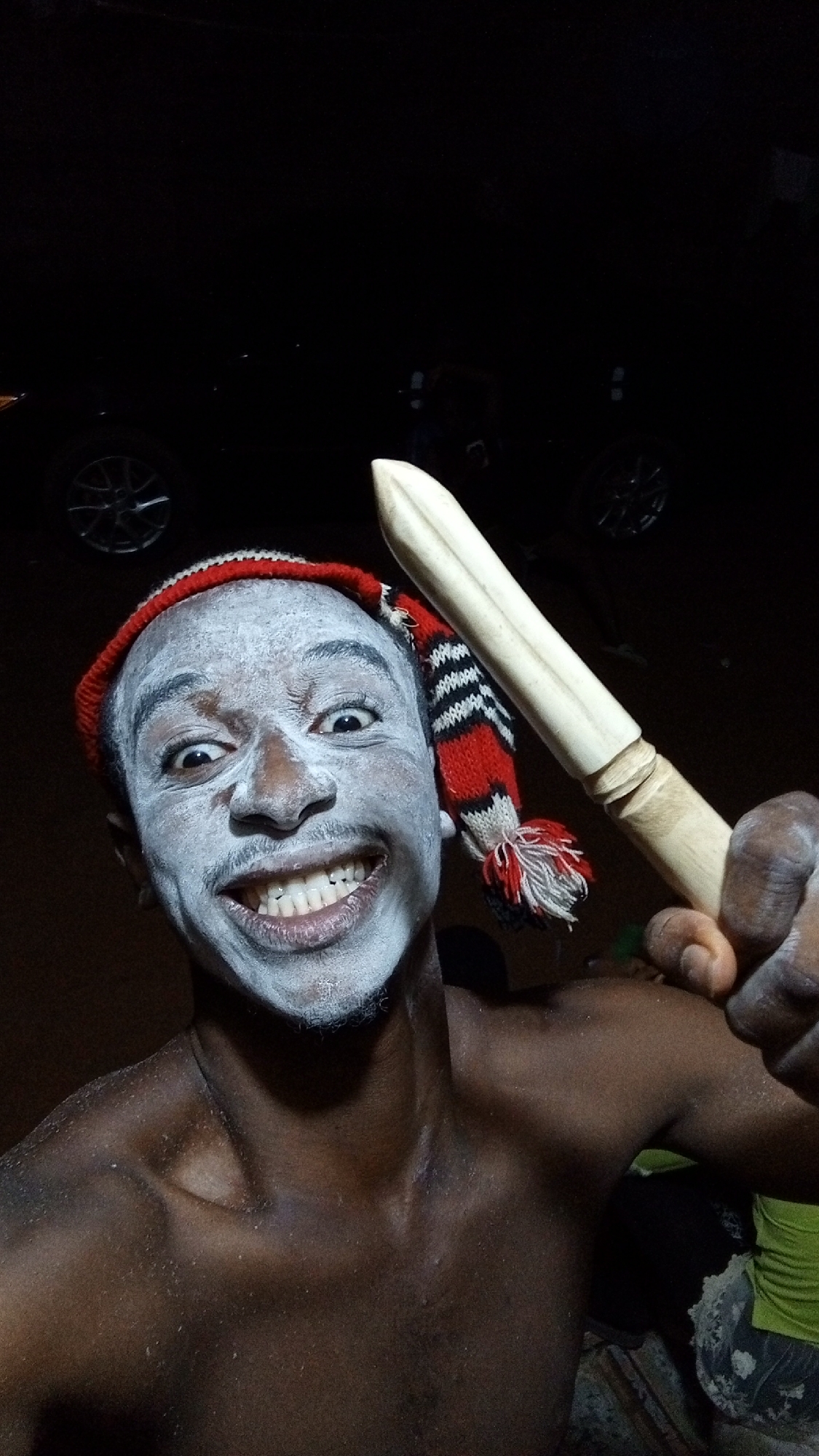 A shirtless Black man with white facial paint, wearing a red and black-striped knit cap, holds a wooden club. The background is dark, revealing a car and hints of other people. The location appears to be outdoors at night, possibly in Onitsha, Nigeria. The man is smiling intensely at the camera.