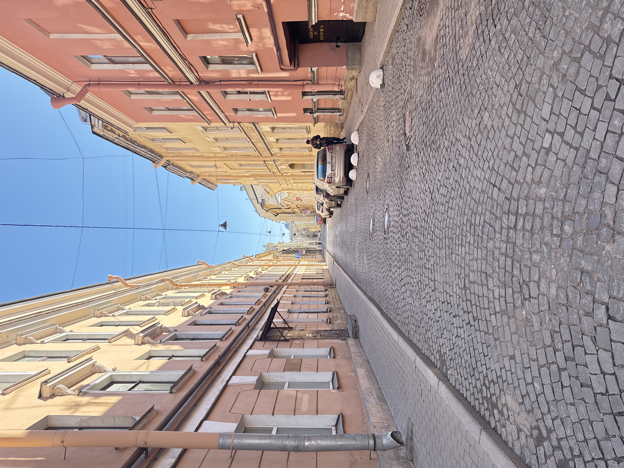 The image shows a narrow street in Saint Petersburg, Russia. The street is paved with cobblestones and lined with buildings on both sides. The buildings are pastel-colored and have multiple windows. A man is standing near a car parked on the street. The sky is clear and blue. The buildings appear to be historic structures. There are power lines running across the street at the top of the buildings. A drainpipe is visible on the lower-left side of the frame. There are manhole covers visible on the street.
