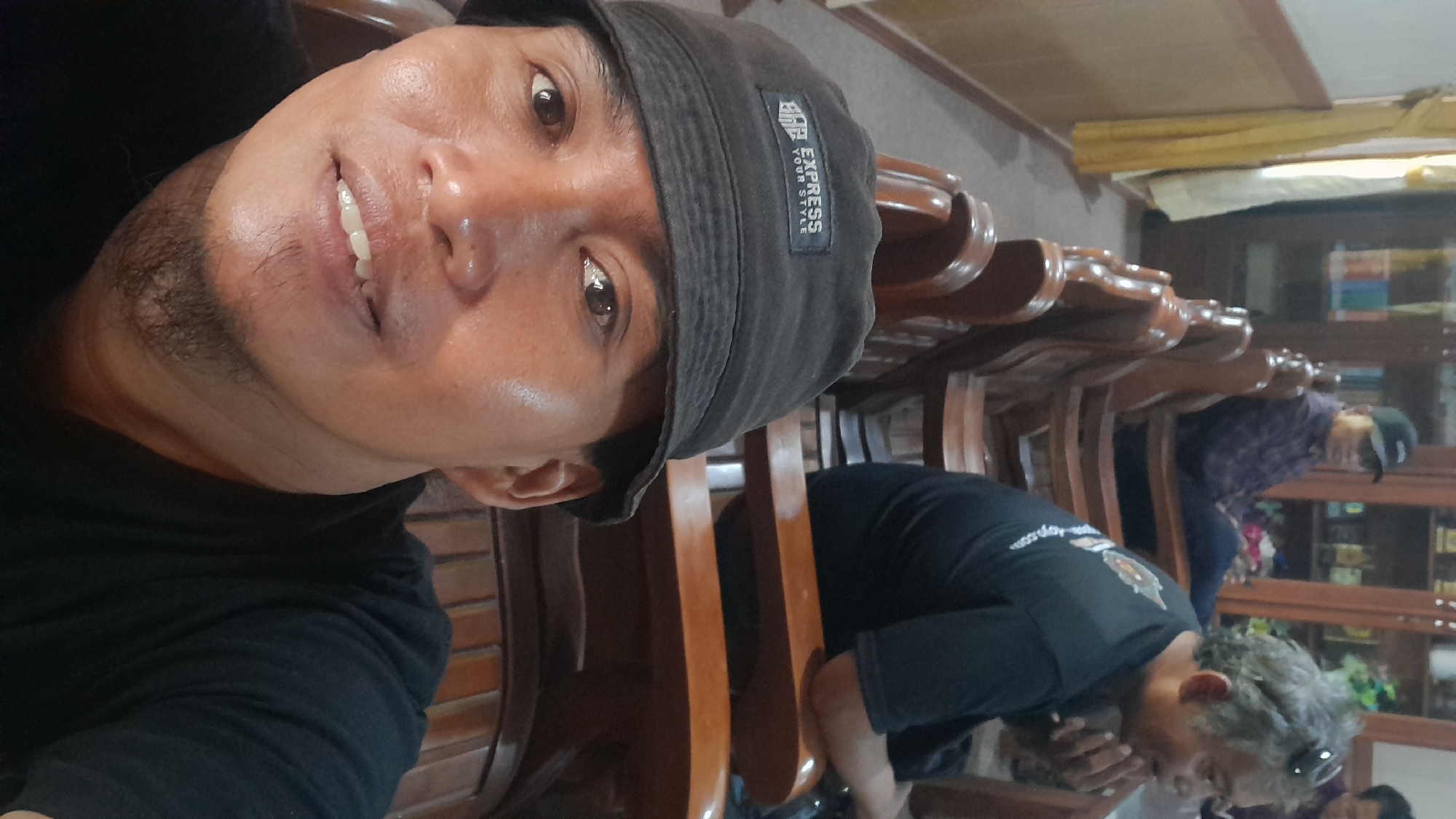 In Blora, Indonesia, a selfie captures a man in a black bucket hat with the text "EXPRESS YOUR STYLE" in white on a black patch. He has fair skin and short, dark hair. His mouth is slightly open, revealing teeth. He has a sparse, dark beard. Behind him, two other men are seated on wooden chairs. The man on the left wears a dark baseball cap and a plaid shirt. The man on the right has gray hair and wears a black t-shirt with a logo on the back. The background features wooden furniture, including chairs and shelves filled with various objects, suggesting an indoor setting, possibly a home or a community hall.