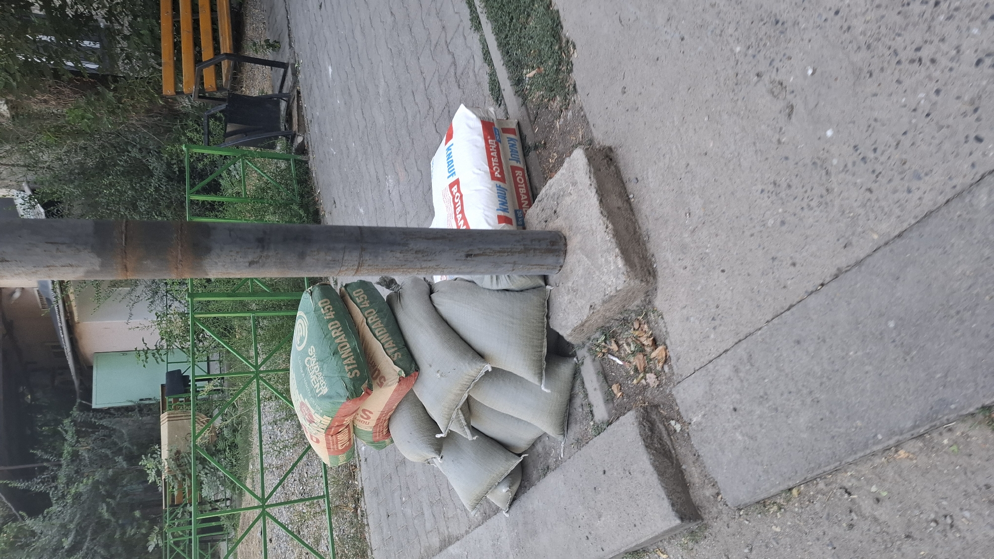 A medium shot captures a pile of construction materials on a paved walkway next to a metal pole. The materials include several bags of cement labeled "STANDARD 450" in green and tan packaging, and a stack of lighter colored, possibly sand-filled, sacks. To the right of the pole, two white bags labeled "KNAUF ROTBAND" are stacked. The background shows a green metal fence, dense foliage, a building with a light-colored facade and a green door, and a wooden park bench. The walkway is made of grey paving stones. The scene appears to be an outdoor urban environment, possibly a residential courtyard or park area in Shymkent, Kazakhstan, given the presence of construction materials and typical urban landscaping. No individuals are present in the frame. The lighting suggests it is daytime.