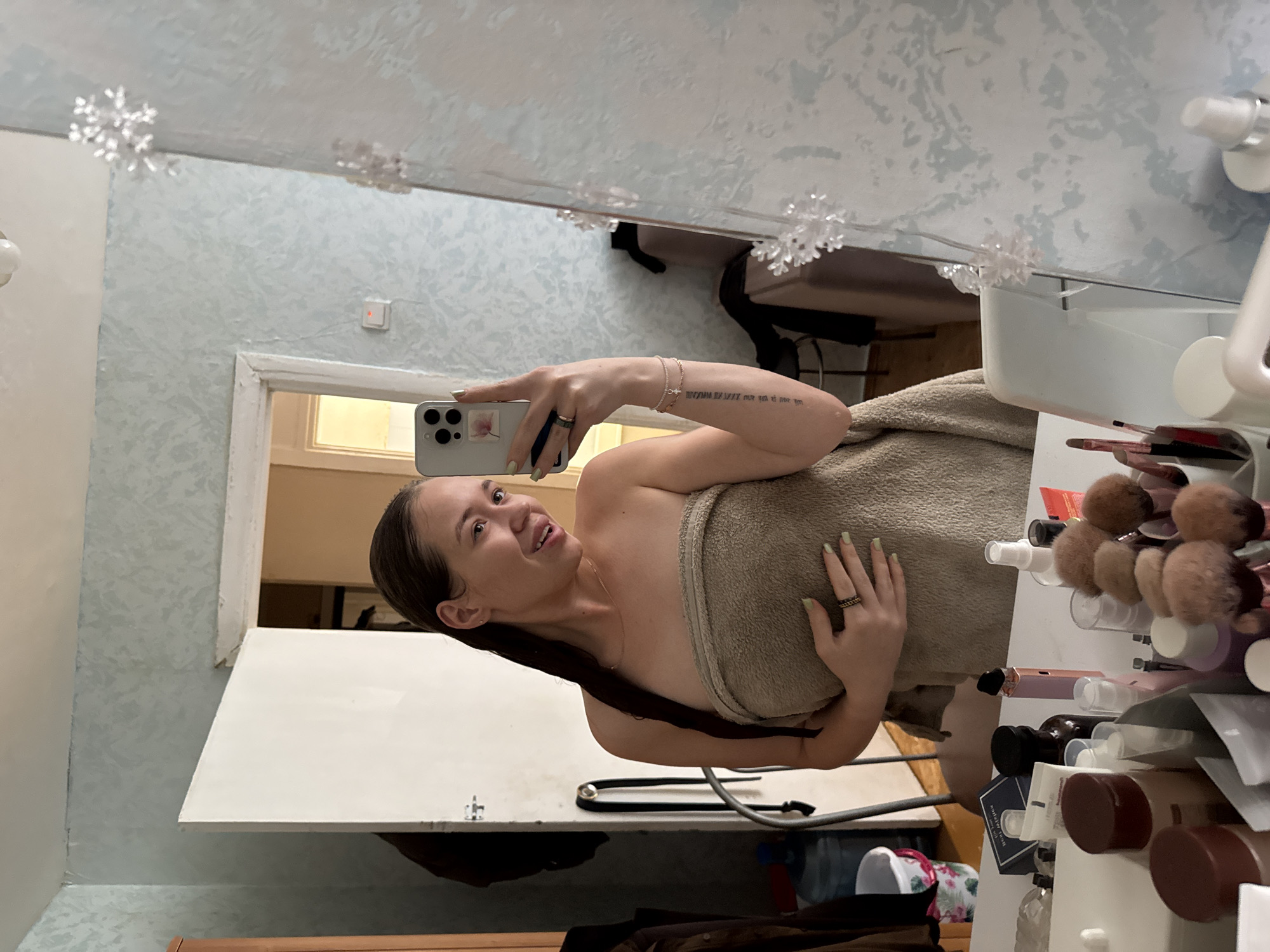 A woman is taking a mirror selfie in a room that appears to be a bathroom or dressing area. She is wrapped in a tan towel, with her wet hair down her back. She is wearing rings on her fingers and bracelets on her wrist. Her phone, which has a floral sticker on the back, is held up to take the photo. On the vanity in front of her, there are various cosmetic items, including bottles, jars, and makeup brushes. The room has light blue wallpaper with a subtle pattern and a white door is visible in the background. The overall impression is that of a private, domestic setting. Based on the information provided, the location is Chaikovsky, Russia.