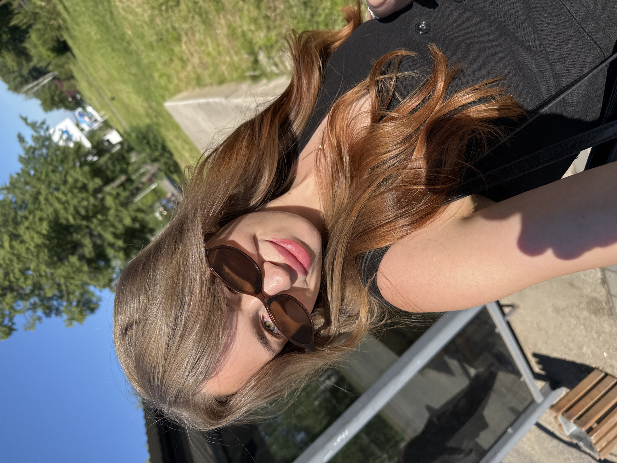 A young woman with long, auburn hair and sunglasses is taking a selfie outdoors on a sunny day. She is wearing a black top. In the background, there are trees, green grass, and a clear blue sky, suggesting a park or a residential area. The overall lighting indicates it is daytime, likely afternoon due to the angle of the shadows. No specific landmarks of Chaikovsky, Russia are visible.