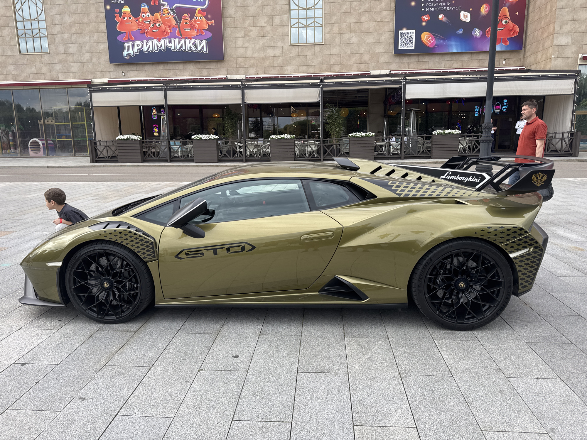 A gold Lamborghini Huracán STO is parked on a paved outdoor area in front of a multi-story building with large windows and awnings. A young boy wearing a dark t-shirt is leaning over the hood of the car, appearing to look at something on the vehicle. To the right of the car, a man in a red polo shirt and blue jeans stands looking towards the building. Above and behind the car, banners with cartoon characters and text are displayed on the building's facade. The environment appears to be a public space in Moscow, Russia, possibly near a shopping center or entertainment venue. The pavement is made of light-colored rectangular tiles.