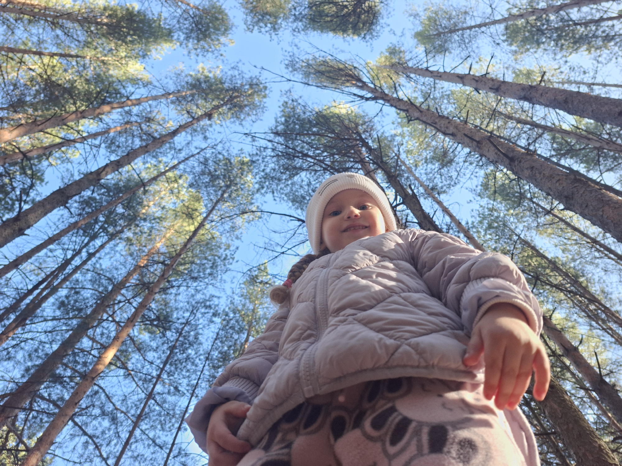 A low-angle shot shows a young child in a white beanie and a light-colored, puffy jacket. The child is positioned in the foreground, looking down with a slight smile. The background features tall trees reaching towards a clear blue sky. The photo was likely taken in a wooded area in the city of Chaikovsky, Russia. The trees are tall and slender, and the sky is bright.