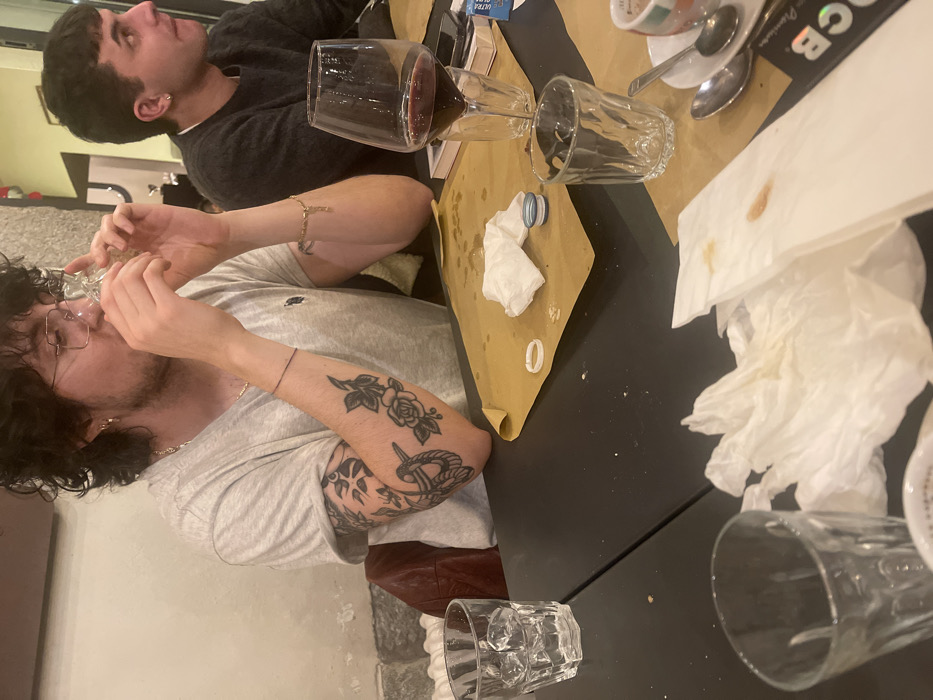 The image depicts an indoor scene, likely a casual restaurant or bar in Sondrio, Italy, featuring two young men seated at a dark table. A brown paper runner or placemat is spread across a portion of the table.
In the foreground, a man with dark, curly hair, glasses, and a beard is actively drinking from a clear glass, which he holds to his mouth. He is wearing a light grey short-sleeved t-shirt, showcasing several tattoos on his left arm, including a prominent rose, an anchor, and other designs. A thin bracelet is visible on his right wrist.
Behind him, another man with short, dark hair and an earring is visible, wearing a dark long-sleeved top. He is looking upwards and to the right, away from the camera.
The table is set with various items suggesting a meal or social gathering. A glass carafe filled with red wine stands next to an empty clear drinking glass. Several crumpled white paper napkins, some stained, are scattered on the table and the paper runner. A small blue bottle cap and a white plastic ring are also present. Further right, a coffee cup resting on a saucer with spoons indicates a finished beverage. A pack of cigarettes with "ULTRA" visible on its blue and white packaging is placed on the table. Another empty clear drinking glass is situated in the bottom right corner of the frame. The scene conveys a relaxed, casual atmosphere, characteristic of an evening outing.