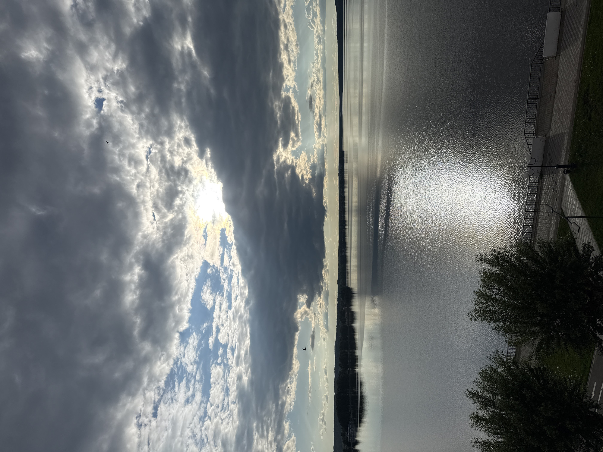 The image presents a dramatic waterscape, likely a large lake or reservoir, captured from an elevated viewpoint. The scene is divided horizontally by a distinct horizon line, with a dynamic sky dominating the upper portion and a broad body of water below.
The sky is a prominent feature, filled with a mix of heavy, dark grey clouds and brighter, more ethereal white clouds. The sun, partially obscured by the dense cloud cover, casts intense light through a break, creating a brilliant area of illumination. This light reflects strongly on the water surface, forming a shimmering path that stretches across the water towards the viewer. Scattered patches of clear blue sky are visible through the cloud breaks, and several small birds can be seen flying.
Below the sky, the vast body of water appears calm, with subtle ripples catching the light. Across the water, a dark, low-lying landmass, possibly a distant forested shore, is visible along the horizon line.
In the foreground, on the right side of the image, there is a paved embankment or promenade. A decorative black metal railing runs along the edge, separating the walkway from the water. Behind the railing, a strip of green grass is visible, and two trees with dark, leafy canopies frame the lower right corner of the shot. A street lamp post is also discernible near the railing.
The lighting suggests either early morning or late afternoon, given the low angle of the sun and its strong reflection. The weather appears partially cloudy to overcast, with the sun breaking through to create a striking visual contrast. The combination of natural elements (water, sky, distant land) with human-made structures (promenade, railing, street light) indicates a developed waterfront area. No visible text is present in the image.