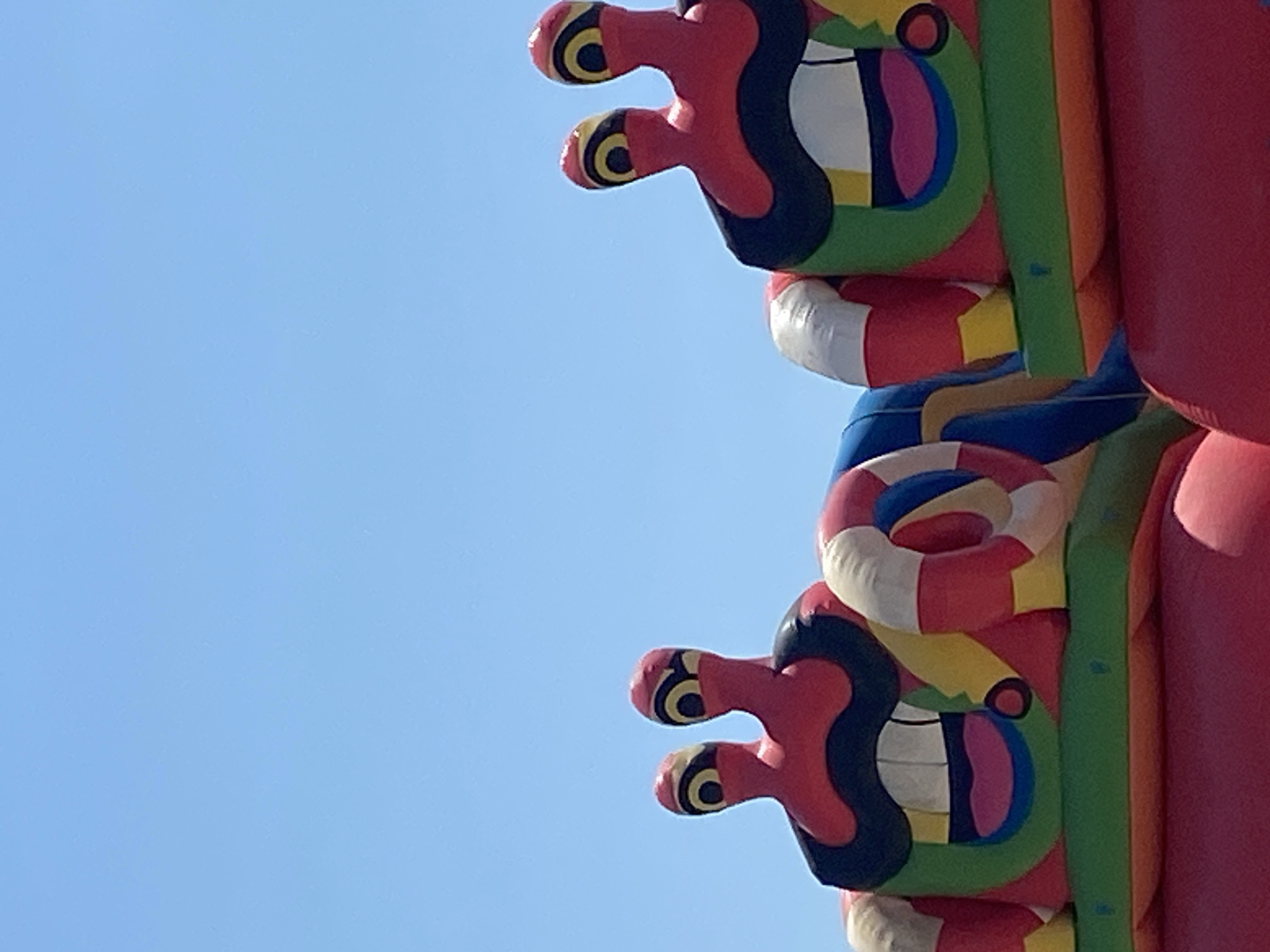 The image shows a portion of a large, inflatable amusement structure, likely a bouncy castle, against a clear blue sky. Two cartoonish crab-like figures with large, red, stalk-like eyes featuring black and yellow markings are visible on top of the structure. These figures have large, black, downturned mouths with white teeth and pink lips. The structure itself is brightly colored with red, green, yellow, blue, and white sections, including what appear to be inflatable rings attached to the sides of the crab figures. The bottom portion of the image shows a large, red inflatable area. The location is identified as Prijevor, Montenegro.