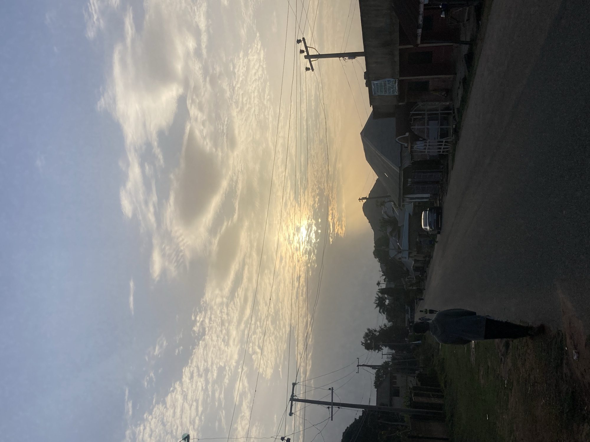 Here's a description of the image:
The photograph depicts a street scene in Jos, Nigeria, taken during the early morning or late afternoon hours, as indicated by the position of the sun. The sky is filled with altocumulus clouds, partially obscuring the sun.
A road runs from the foreground to the center of the image. Utility poles with power lines run along the left side of the road. Buildings are present on the right side of the road. A person is walking alongside the road in the foreground.