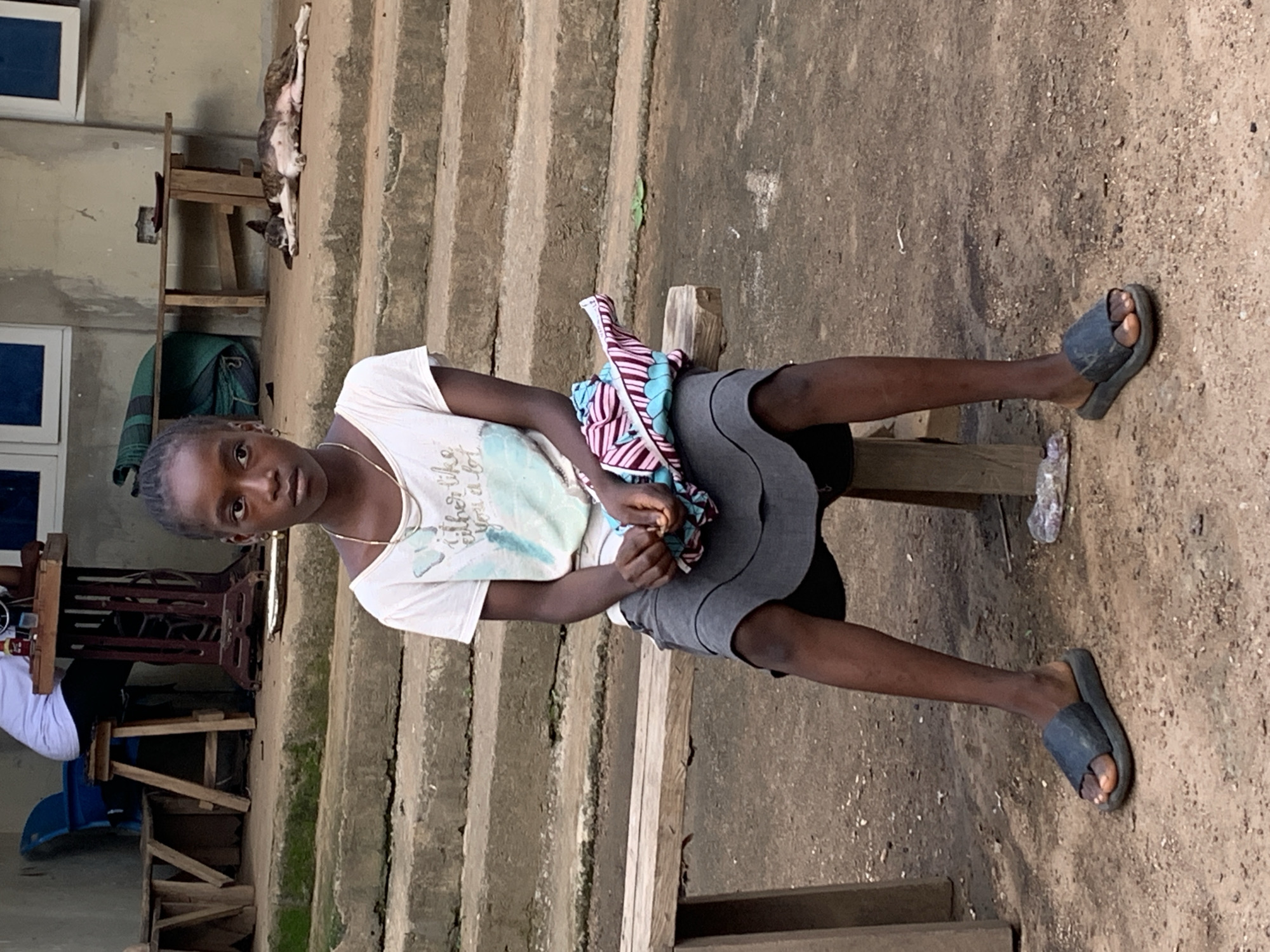 A young Black female sits on a wooden bench, looking directly at the camera. She has dark skin, dark braided hair pulled into a bun, and is wearing a white t-shirt with a graphic design, a dark grey skirt, and dark blue slides. She is holding a folded, patterned cloth in her lap. The background shows a concrete staircase, a building with windows, and a wooden table where a dog is lying down. The scene appears to be outdoors, possibly in a residential area of Akwanga, Nigeria.