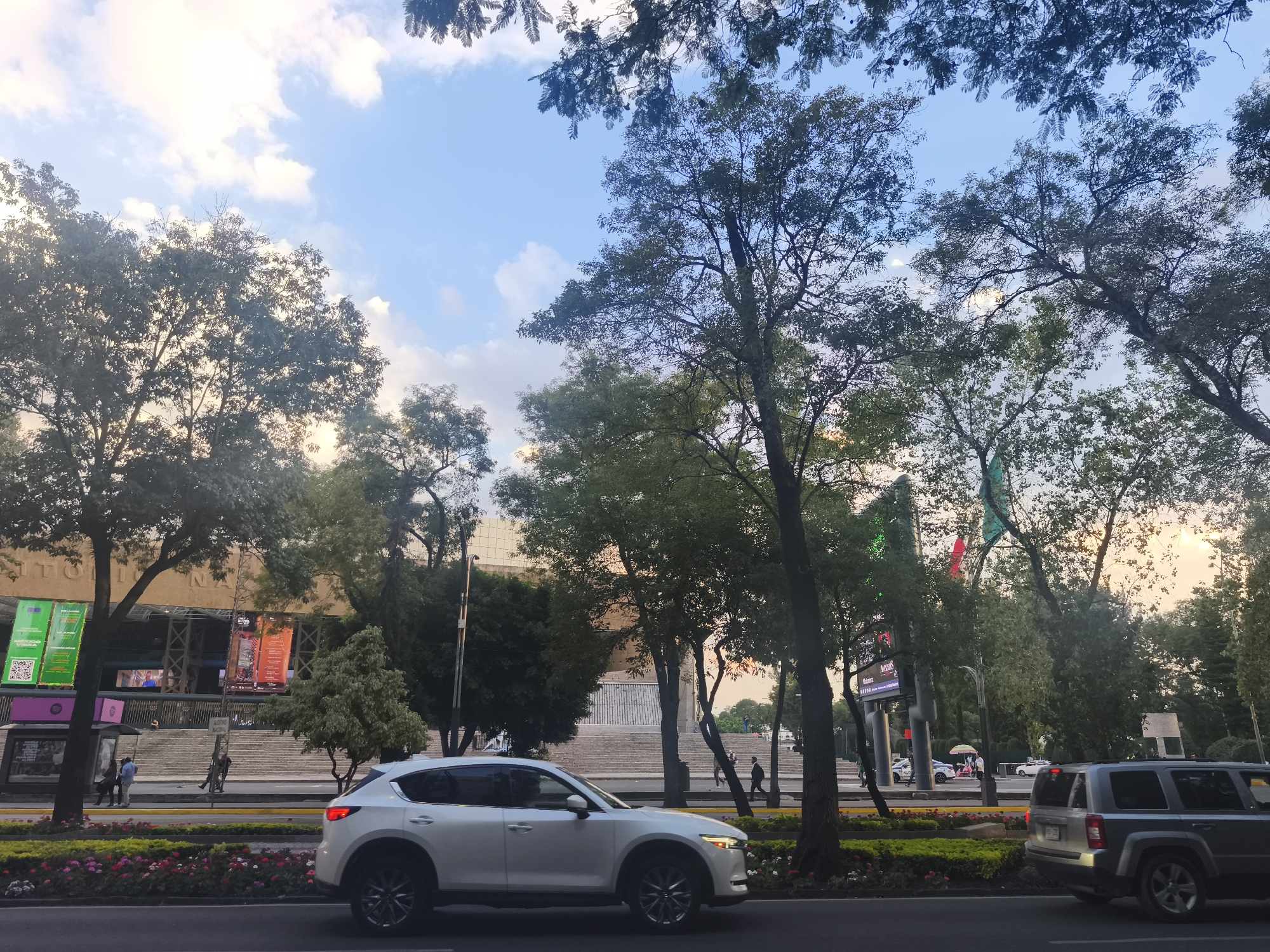 This image captures a vibrant street scene in Mexico City during what appears to be late afternoon or early evening, evidenced by the soft, warm light filtering through the trees and the varied hues of the sky.
The scene is dominated by a wide, tree-lined street with a median strip planted with colorful flowers. A white SUV, possibly a Mazda CX-5, is prominently featured in the foreground, traveling from left to right. Further down the road, a dark grey SUV, resembling a Jeep Patriot, is partially visible, moving away from the viewer.
In the background stands a large, modernist building with expansive, monumental steps leading to its entrance. Visible on the building's facade is the text "AUDITORIO NACIONAL," identifying it as a significant public venue. Several people are seen walking on the steps and sidewalks.
On the left side, near the building, a bus stop or shelter is present, featuring a distinct purple Spotify logo. Bright promotional banners hang near the building's entrance; one is green with QR codes and text, while another is reddish-orange, likely advertising an event.
The street is heavily shaded by numerous tall trees with dense, green foliage, contributing to a park-like atmosphere within the urban setting. Through the trees on the right, a traffic light and a partial view of a flag with green, white, and red stripes (likely the Mexican flag) are visible. The sky above is a mix of light blue with scattered white and creamy clouds, indicating fair weather. The overall impression is a bustling urban environment integrating nature with prominent architectural landmarks.