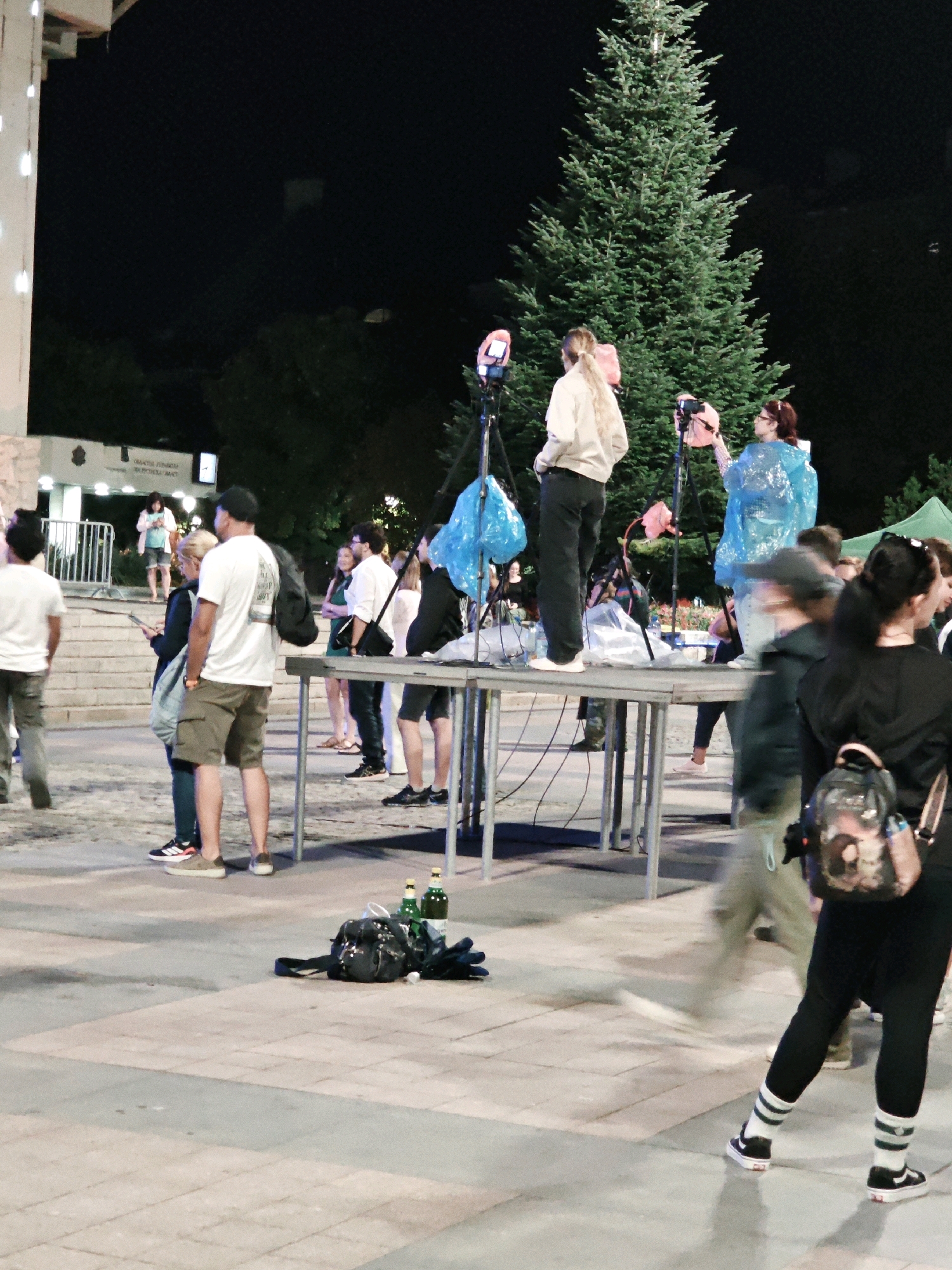 In an outdoor urban setting at night, a group of people are gathered around a low table with equipment. A tall evergreen tree is visible in the background, along with a building facade and streetlights. On the table, there are several pink lights on tripods, two blue plastic bags, and various other items that are difficult to discern.
Several individuals are standing on or near the table. One person, wearing a light-colored jacket and black pants, is standing on the table and facing away from the camera. Another person, wearing glasses and a blue poncho, is also on the table, appearing to operate a camera on a tripod. To the left, two men in casual attire, one in a white t-shirt and shorts and the other in a white t-shirt and grey pants, are observing the scene. A woman in a black top and patterned leggings is walking past in the foreground, her back to the camera.
On the ground near the table, there is a black backpack and two green bottles. The ground is paved with interlocking stones. The lighting suggests it is evening or night, with artificial lights illuminating the area. The overall scene appears to be related to an outdoor event or performance setup.