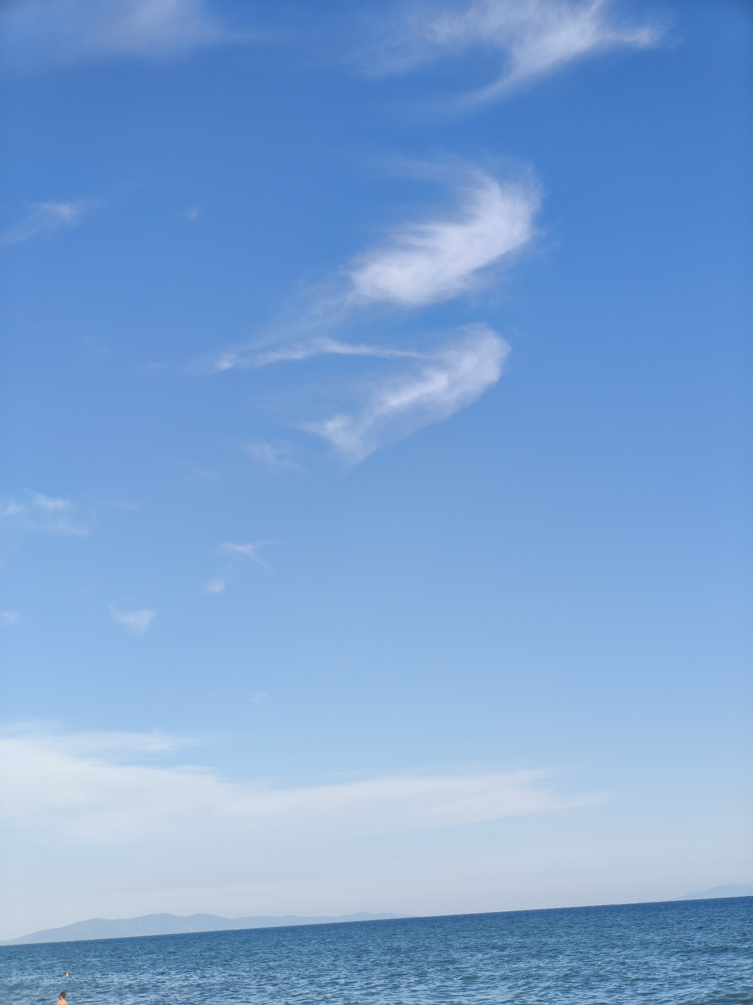 This is a vertical, eye-level shot of a serene seascape at Castiglione della Pescaia, Italy. The image is dominated by a vast expanse of clear blue sky, adorned with wispy white clouds that form a shape resembling a stylized bird or a crescent moon. In the distance, the calm, blue sea stretches towards a hazy horizon, where faint, undulating hills are visible. The water is slightly rippled, catching the light and creating a gentle shimmer. A small portion of the scene at the bottom left reveals a hint of a beach with two people visible in the water, one wearing an orange float. The overall atmosphere is one of tranquility and natural beauty.