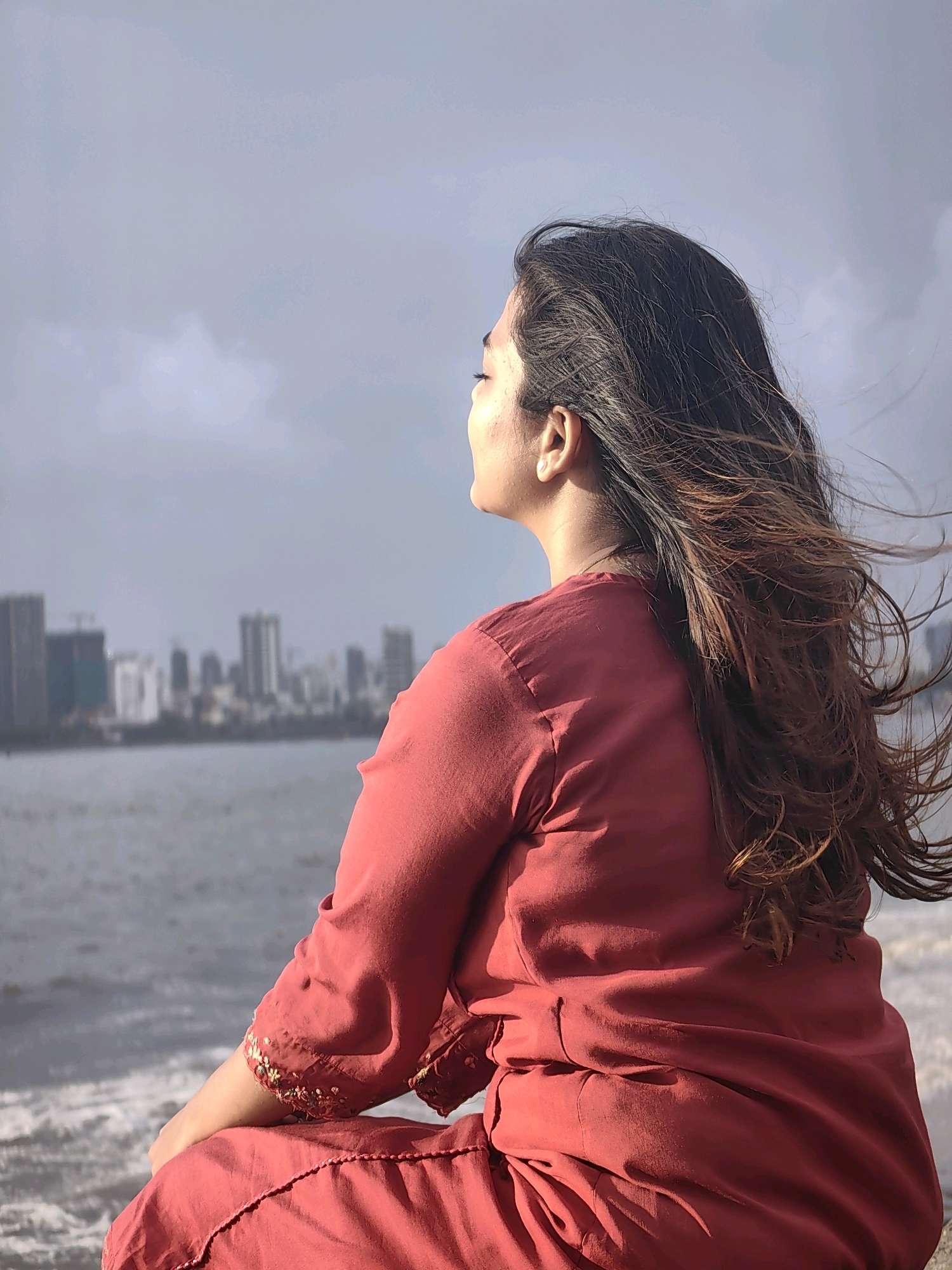 A young woman with long, dark, wind-swept hair is seated on a ledge, facing away from the camera and looking out at a body of water with a city skyline in the distance. She is wearing a reddish-pink kurta, a traditional Indian tunic, with embroidered sleeves. The water appears to be choppy, with waves breaking gently. The city skyline consists of several tall buildings, indicating an urban setting. The sky is overcast with a mix of clouds and patches of sunlight. The overall impression is one of contemplation and solitude. The image appears to have been taken at Marine Drive in Mumbai, India, given the characteristic skyline and the waterfront setting.
