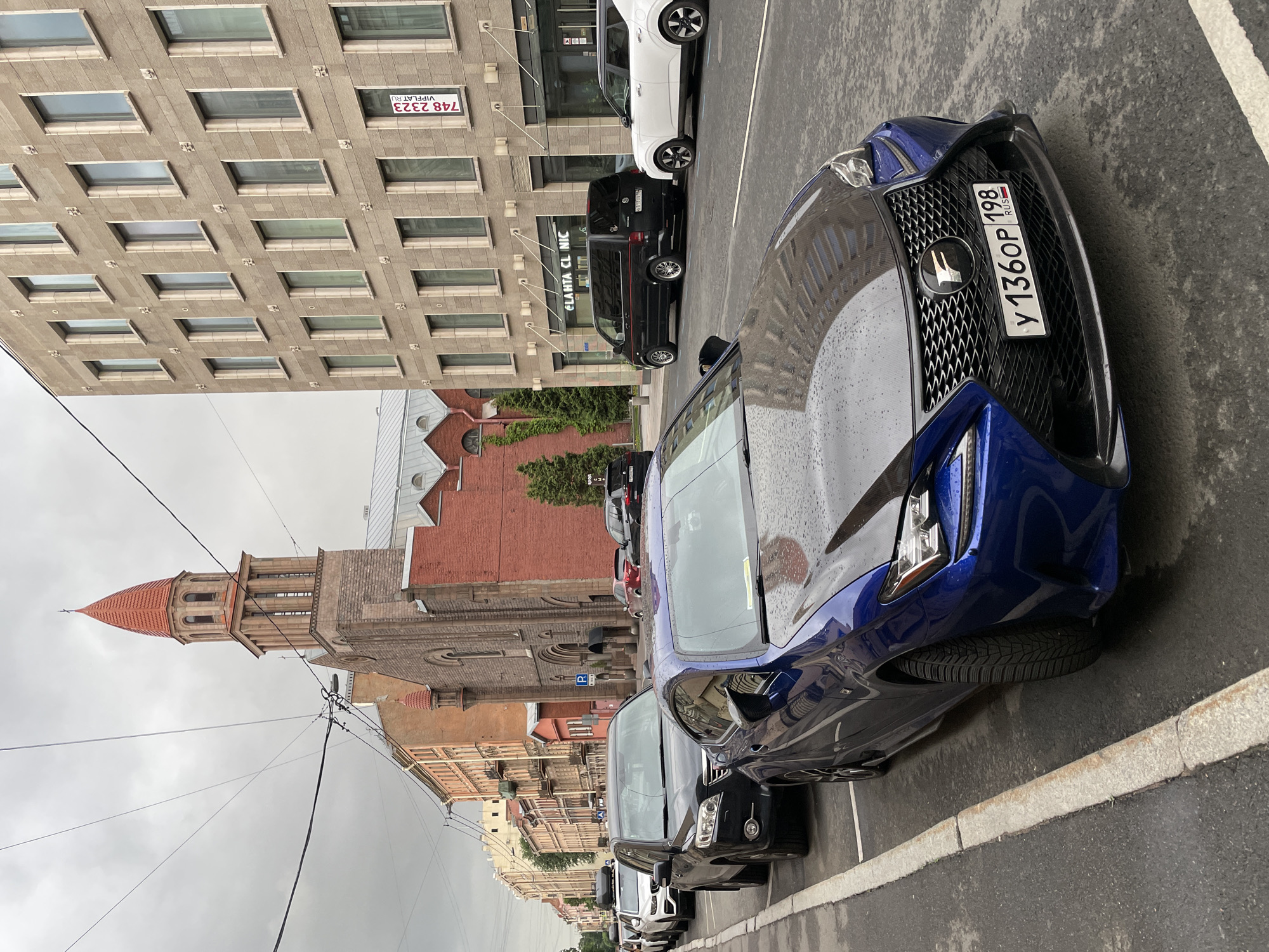 The image depicts a street scene in Saint Petersburg, Russia, on a cloudy day. In the foreground, a blue Lexus RC F is parked, its hood reflecting the surroundings. Beyond it, a line of cars is parked along the side of the road, including a black SUV, a white SUV, and several other vehicles. A tall, red-roofed church with stone accents stands prominently on the right side of the street. Adjacent to the church is a modern, beige building with many windows and a sign that reads "CLAHTA CLINIC". Another building, appearing older with orange facades, lines the left side of the street. Overhead, electrical wires crisscross the sky, and the wet asphalt suggests recent rain. No people are visible in the image.