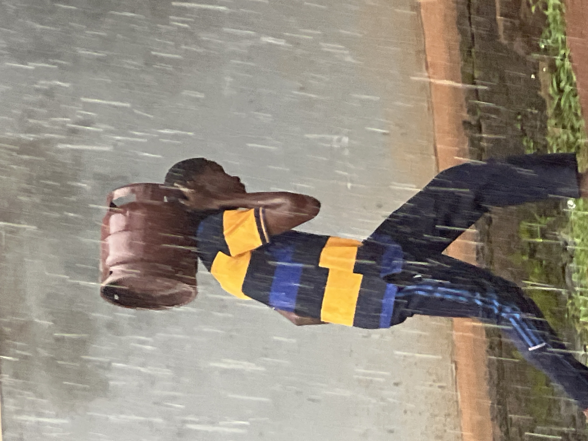 The image shows a person in Nkpor, Nigeria, carrying a brown barrel over their shoulder while walking in the rain. The person is wearing a blue and yellow striped t-shirt and dark pants with blue stripes. The rain is heavy, with streaks of water visible in the air and on the ground. The setting appears to be a street or pathway, with a brick wall and greenery visible on the right side of the image. The overall impression is one of a busy person on a mission, undeterred by the inclement weather. The time of day is not discernible due to the overcast conditions and the rain. There is no visible text in the image.