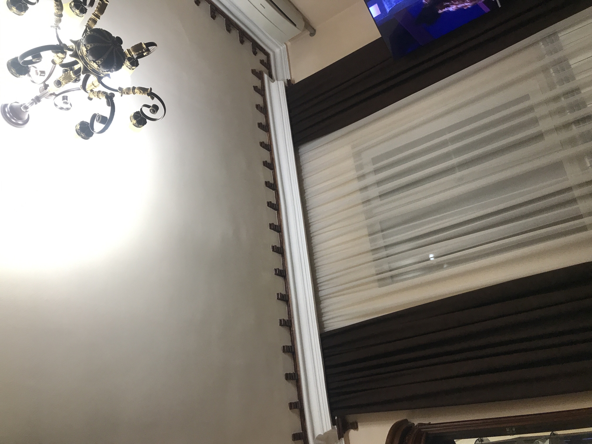 The image shows an interior view of a room. Visible are a chandelier, a wall with decorative molding, window blinds, and a television screen. The chandelier is ornate, with multiple bulbs and scrolling arms. The wall is painted a light color and has decorative molding along the top edge. The blinds are horizontal and layered, providing partial light control. The television screen displays a blurry image, but it's unclear what it depicts. The location is not discernible from the image. There are no people visible in the image.