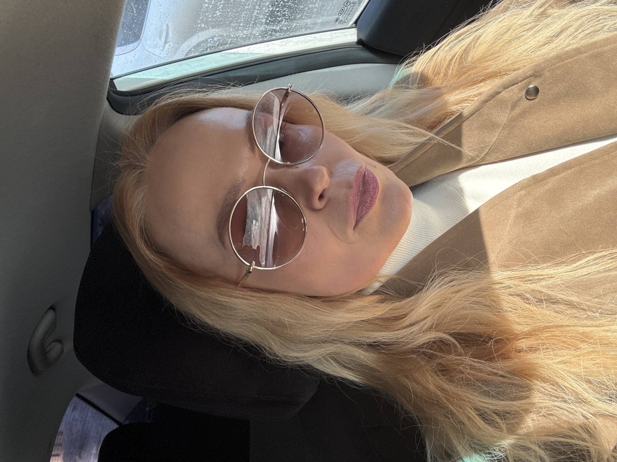 A woman with blond hair and round sunglasses is resting in the back seat of a car. She is leaning against a black headrest and wearing a tan jacket over a white top. The background shows the car's window with what appears to be rain or moisture on the glass. The location is unspecified in the prompt; Chaikovsky, Russia is not discernible from the image.